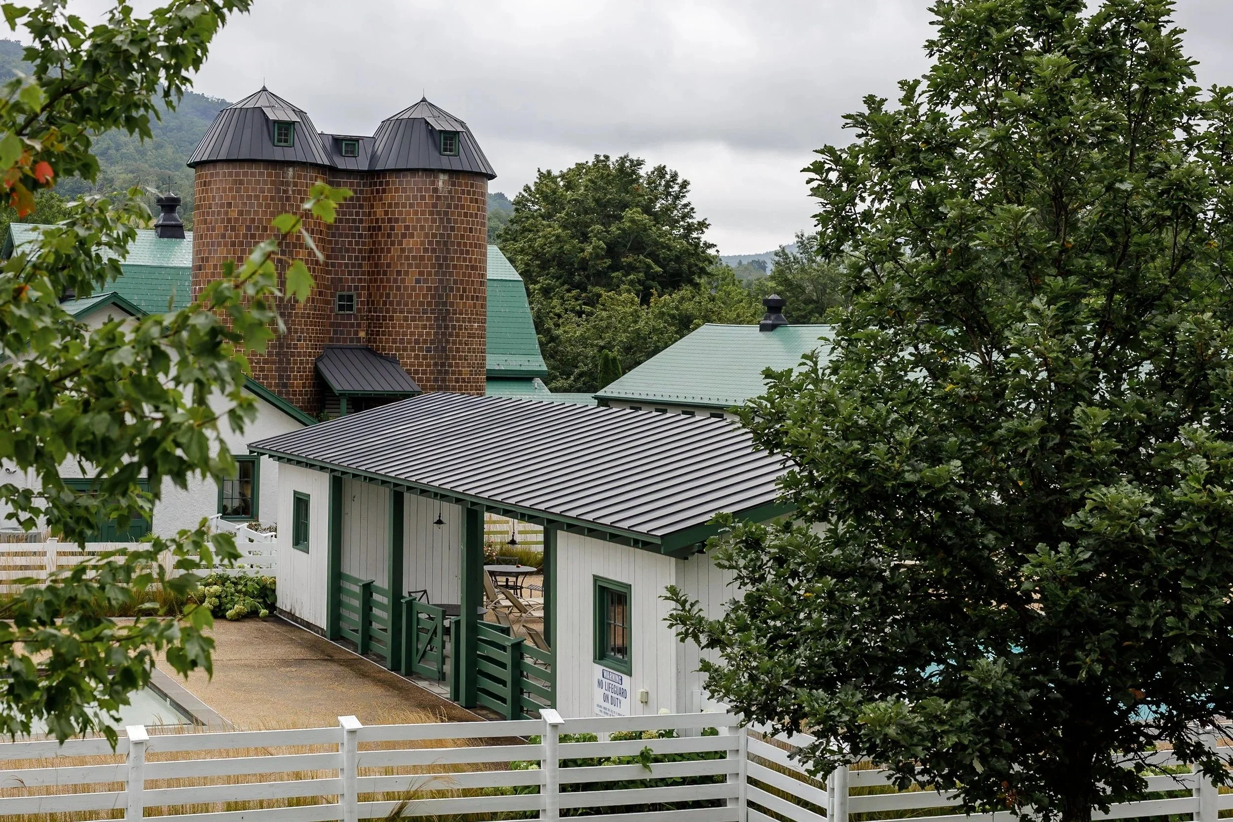 Residential buildings with gray metal roofs, surrounded by trees and green mountainous landscape in the background, under an overcast sky.