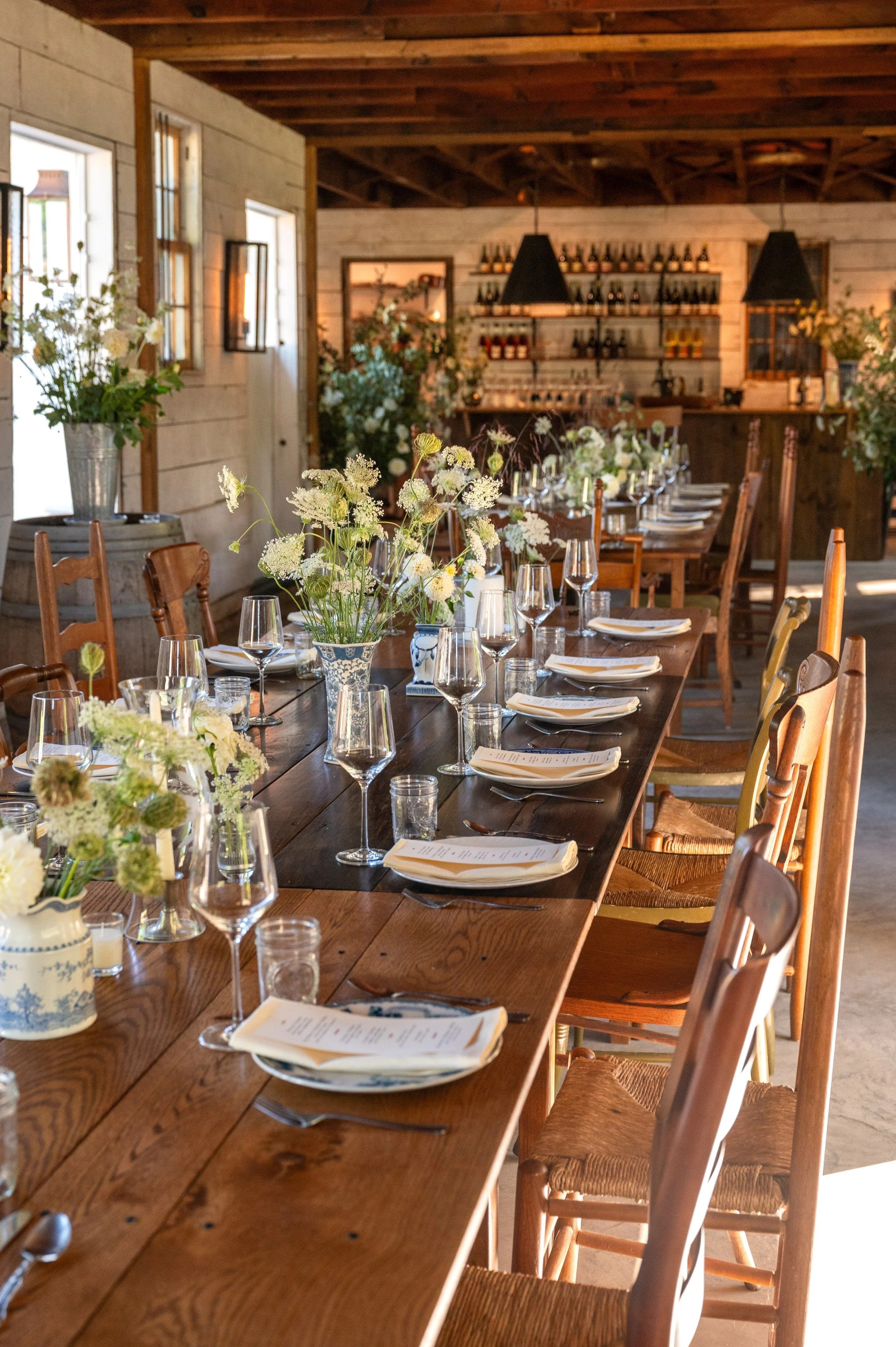 A rustic indoor dining setup with a long wooden table, decorated with white flowers in vases, filled with wine glasses, plates, and napkins. The background features a wall with shelves lined with bottles, and evidence of a cozy, warm ambiance with natural light coming through the windows.
