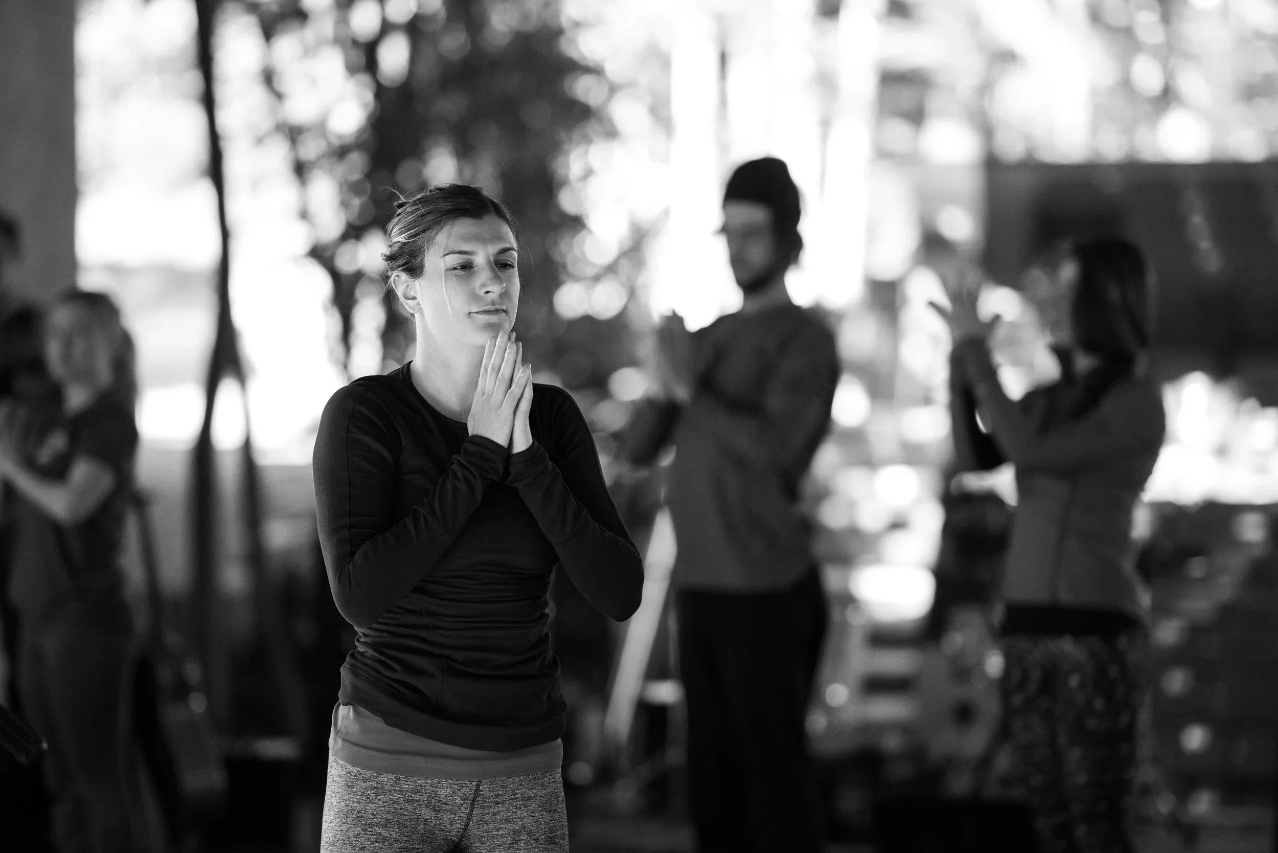 A woman in athletic clothing practicing yoga or meditation indoors, with her hands pressed together near her face, surrounded by others in similar poses, creating a calm and focused atmosphere.