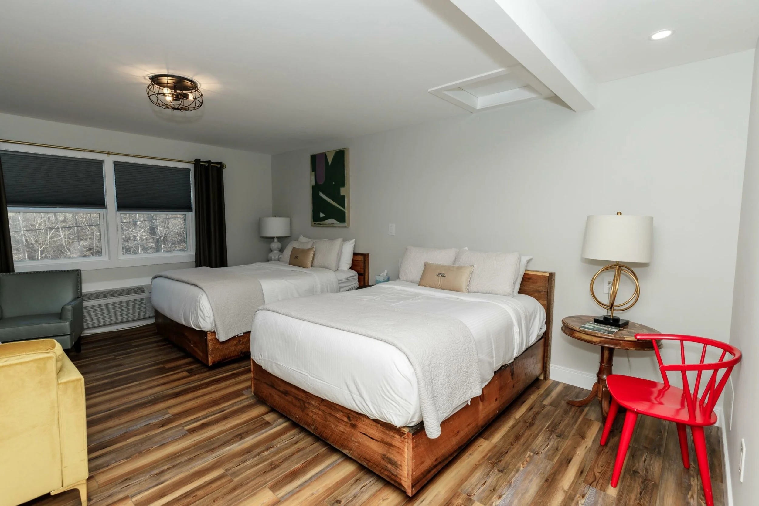 A bedroom with two beds with white bedding, a nightstand with a lamp, a painting on the wall, large windows with black blinds, a green armchair, and red chairs around a small wooden table. The room has wood flooring and a ceiling light fixture.