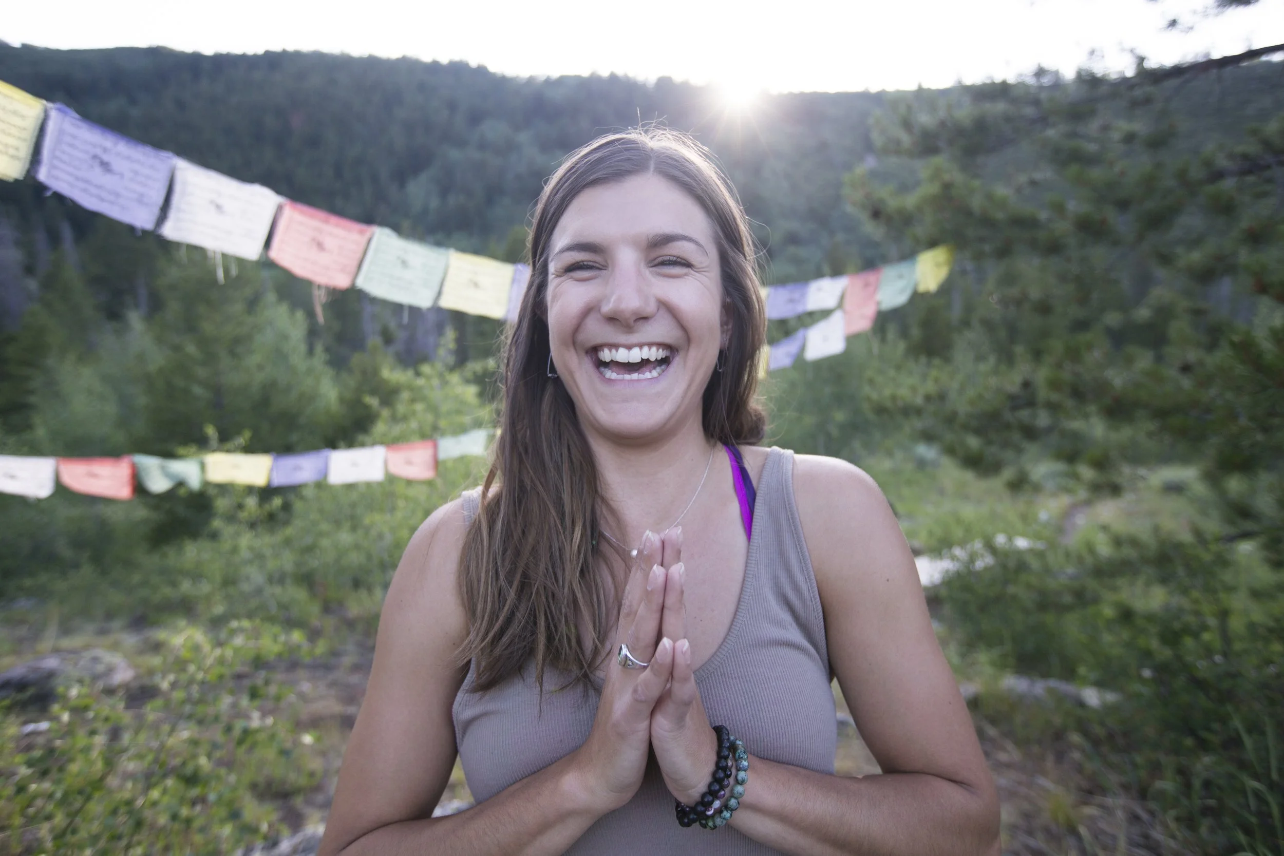 A woman smiling and doing a meditative pose with hands in prayer position outdoors, with prayer flags and a mountain landscape in the background.