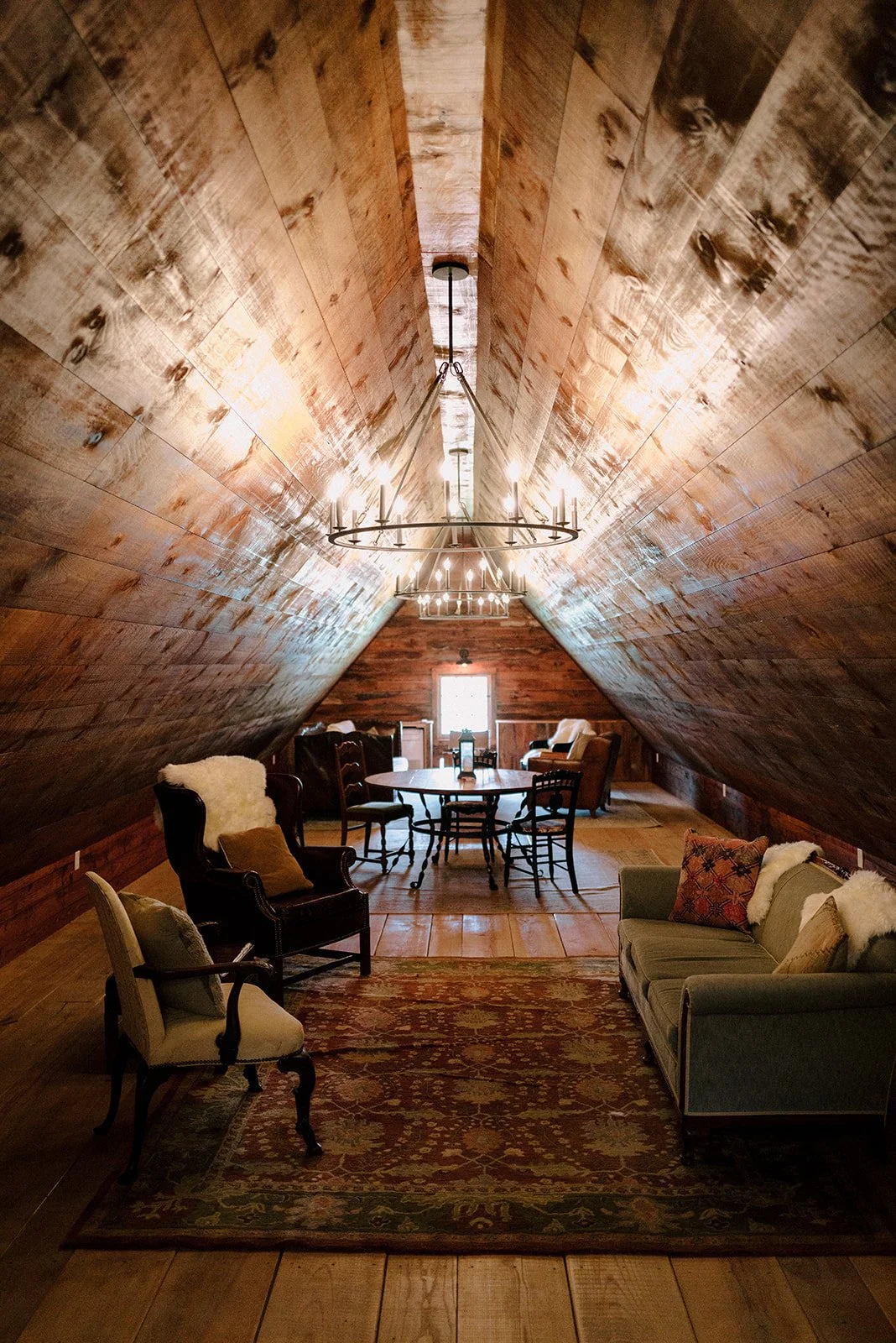 Attic room with wooden sloped ceiling, chandelier lighting, and vintage-style furniture including sofas, armchairs, and a round dining table.