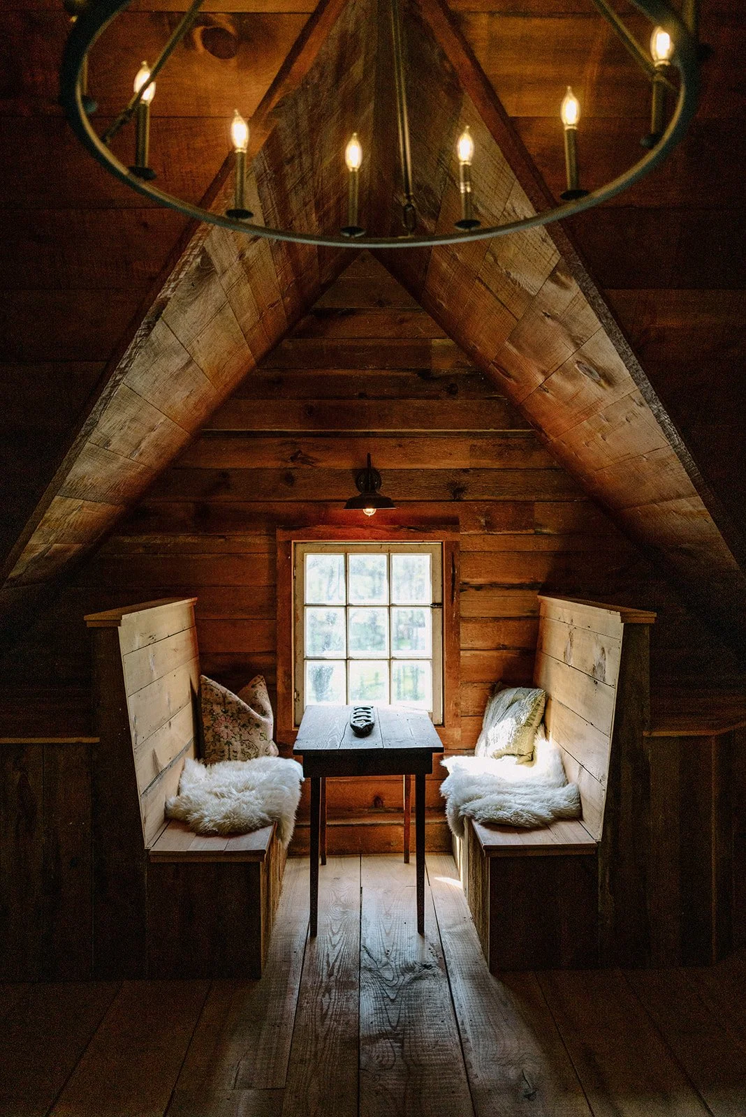 Cozy nook with two wooden benches with pillows and fur throws, positioned on either side of a small table, in a wood-paneled attic space with a window and a chandelier overhead.