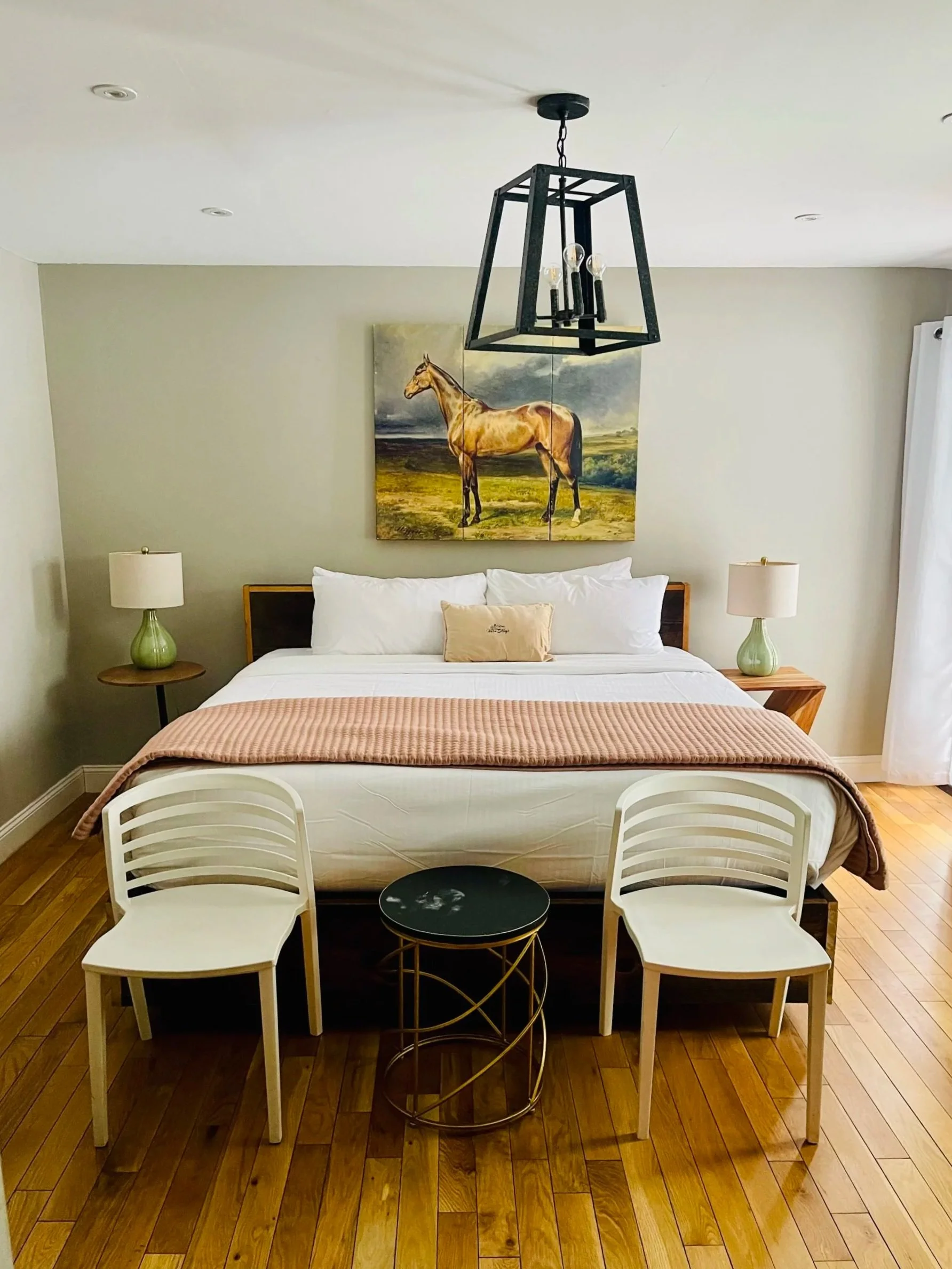 A bedroom with a bed, beige walls, two nightstands with lamps, a painting of a horse above the bed, and a black hanging light fixture. There are two white chairs and a small black round table in front of the bed on a wood floor.