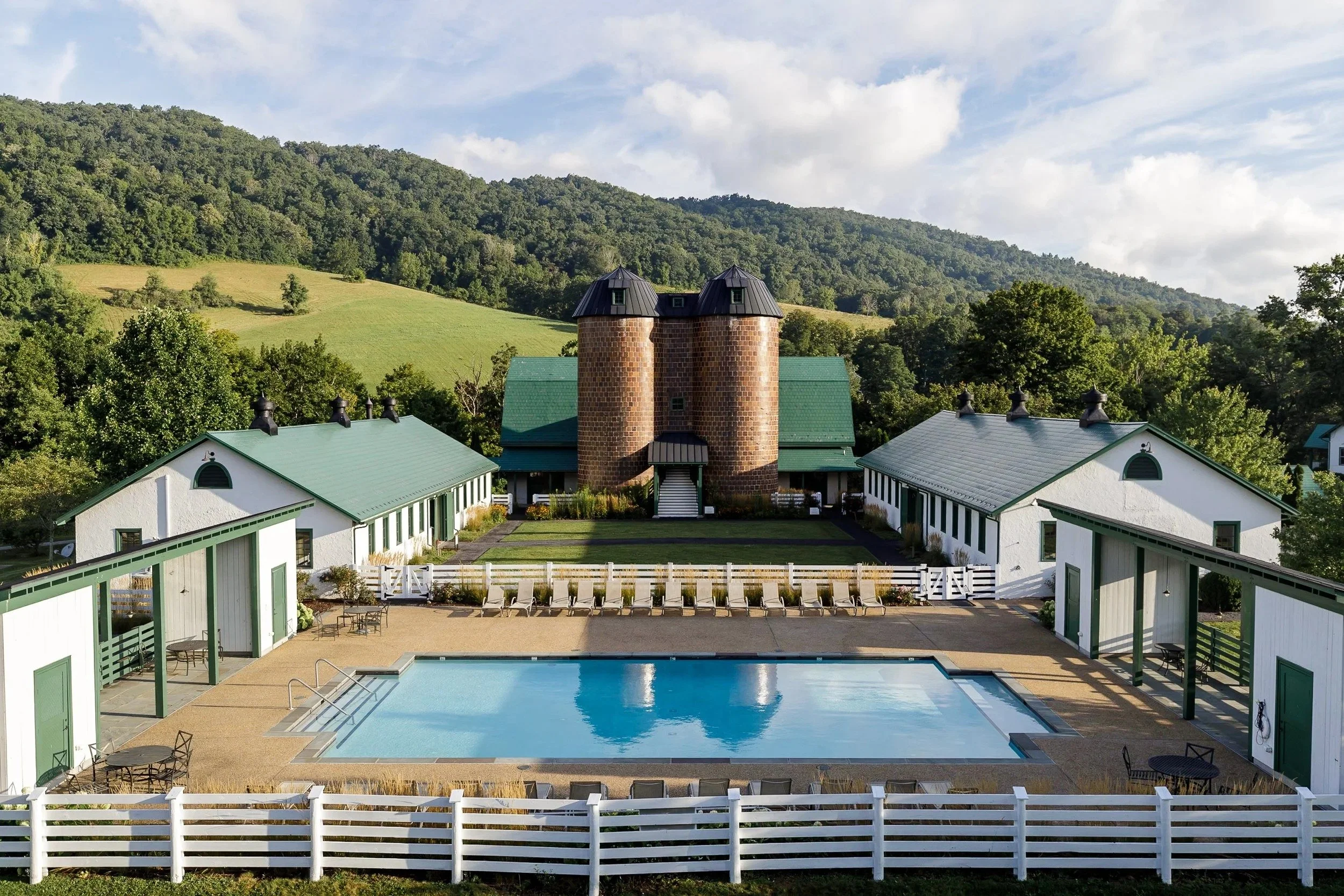 A large outdoor swimming pool with lounge chairs, surrounded by a white fence, with a hotel or clubhouse building and lush green hills in the background.