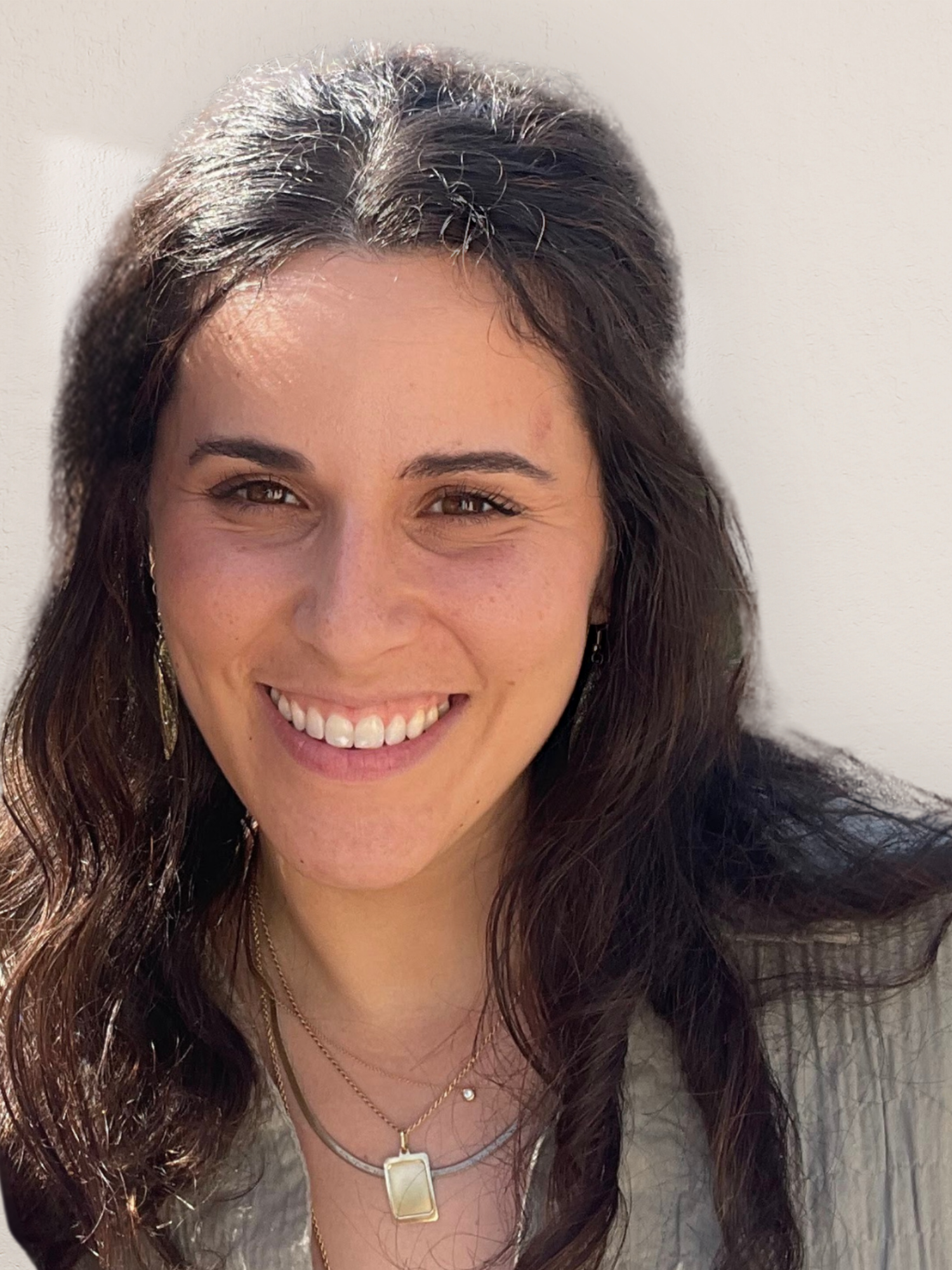 Smiling woman with long, dark, wavy hair and brown eyes, wearing jewelry, against a plain light-colored background.