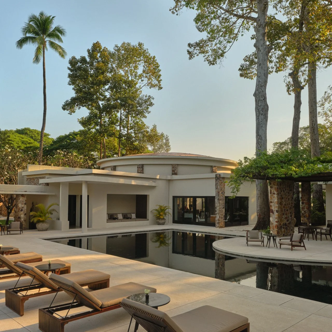 Modern house with outdoor pool, surrounded by trees, with lounge chairs in the foreground and a clear sky above.