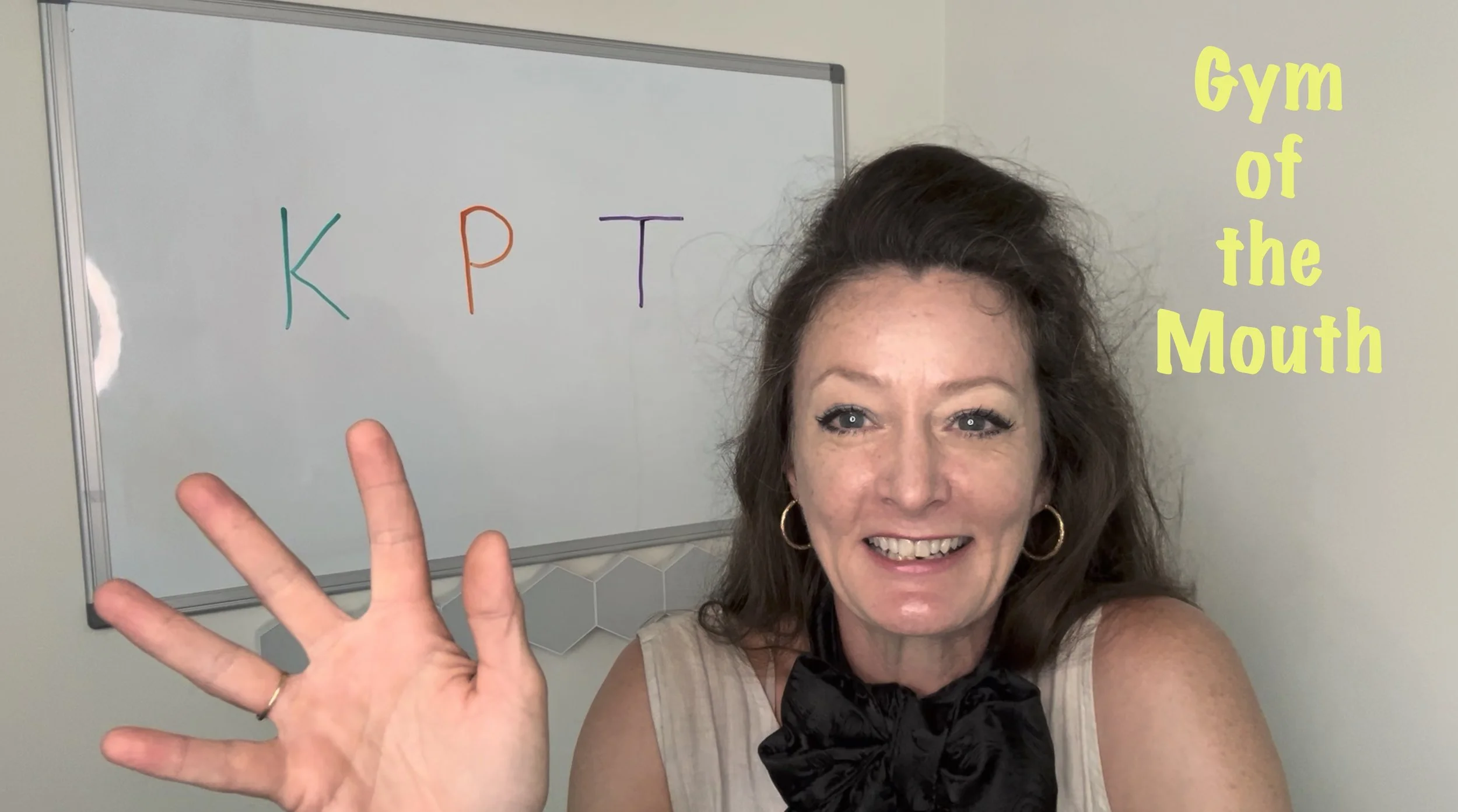 A woman smiling and holding up four fingers in front of a whiteboard with colorful letters 'K', 'P', and 'T'. Text on the right side says 'Gym of the Mouth'.