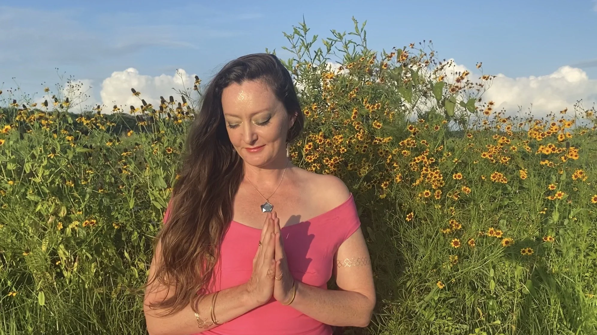 A woman with long brown hair, wearing a pink off-the-shoulder top, stands with her eyes closed and hands pressed together in a prayer position, in a field of yellow wildflowers under a blue sky with some clouds.