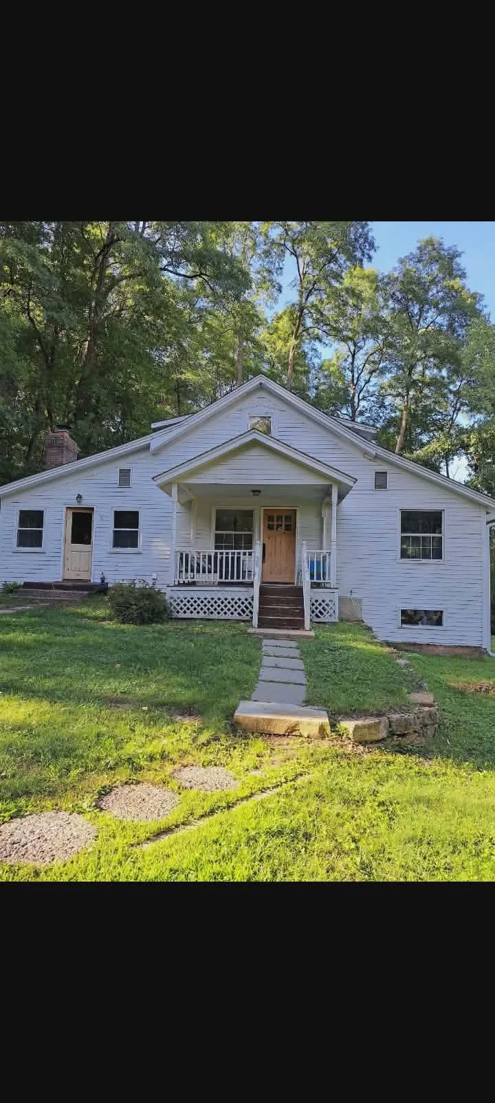 White house with a central front porch, wooden door, and a pathway leading to it, surrounded by green grass and trees. Before picture of exterior house painting project.