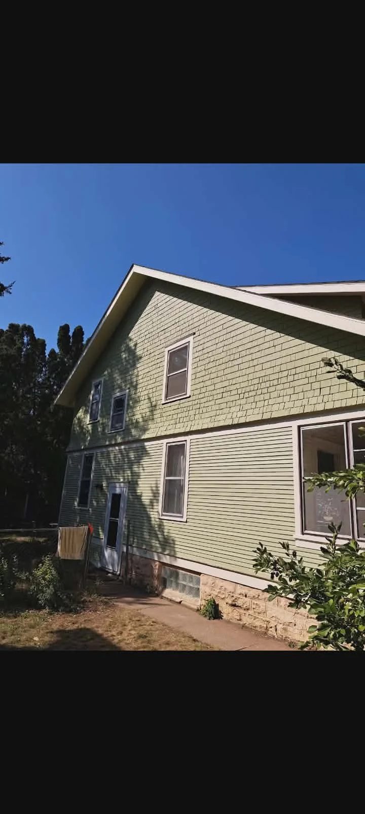 A two-story house with light green exterior siding and multiple windows under a clear blue sky. The house has a sloped roof and a small yard with a sidewalk and some plants.