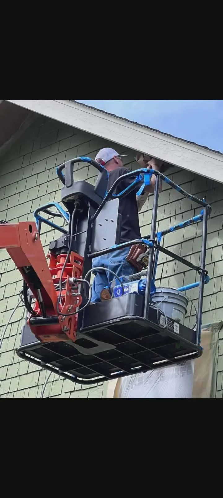 Person wearing a white cap and gray shirt working on the side of a house from a lift platform.