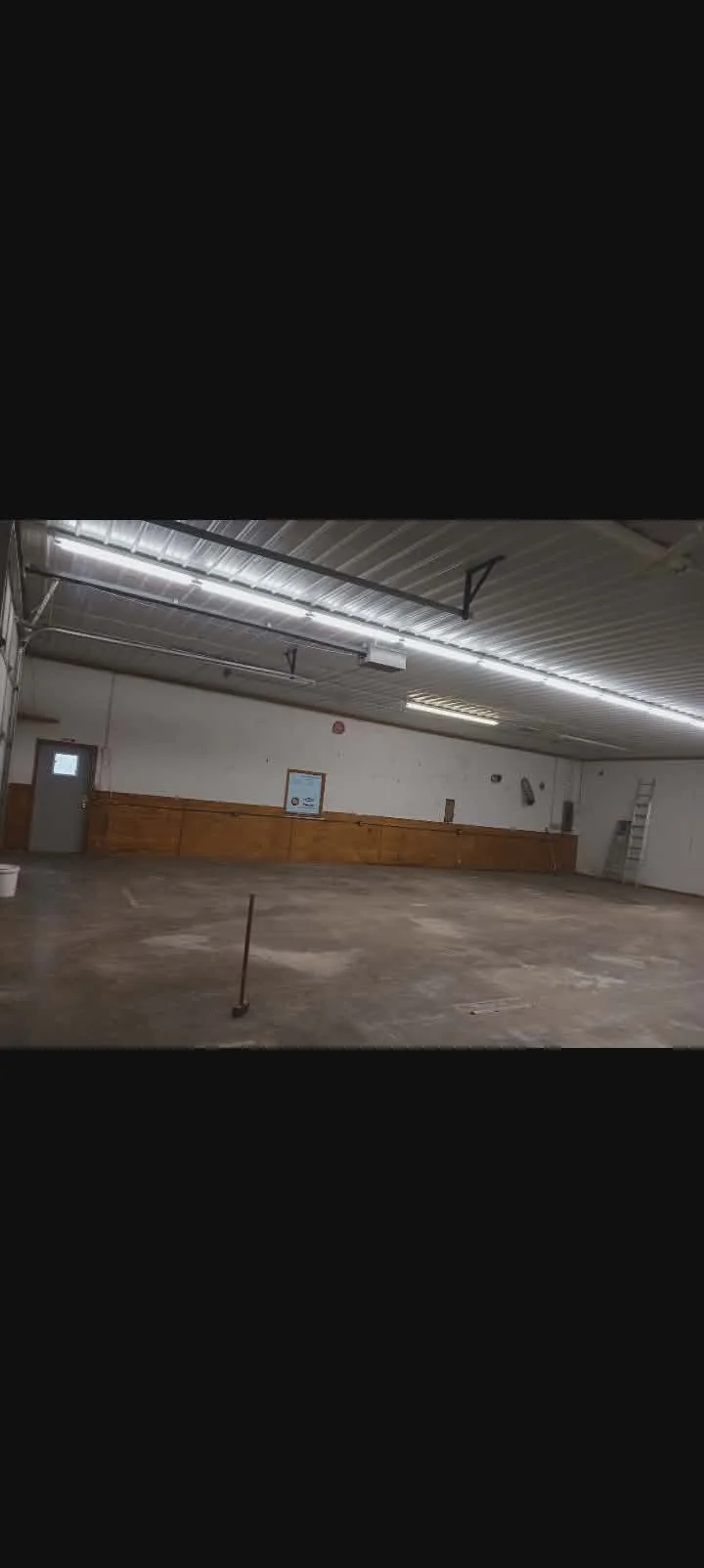 Empty indoor warehouse or garage with fluorescent lighting, a ladder in the corner, and a long wooden counter along the wall. Bessemer Small Engine remodel. Before paint project picture 