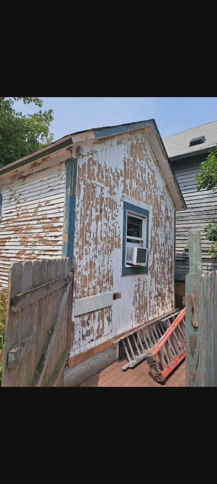Old house with peeling paint on the exterior walls, a window with an air conditioning unit, and a wooden fence in the foreground. Before painting project picture 
