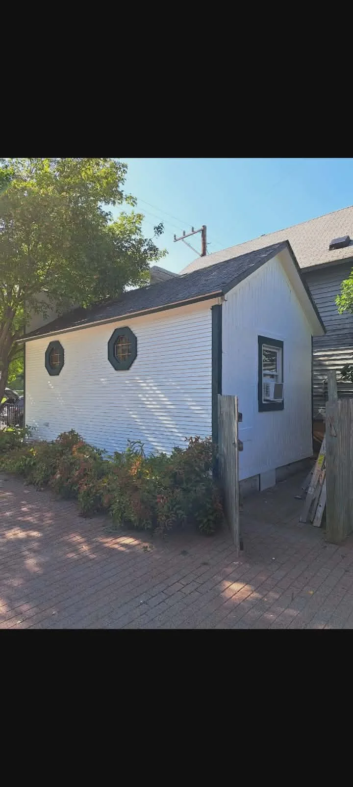 A small white building with two octagonal windows, a black window frame, and a window air conditioning unit, surrounded by a brick sidewalk and some bushes, with a large tree and a neighboring house in the background. After painting project picture 