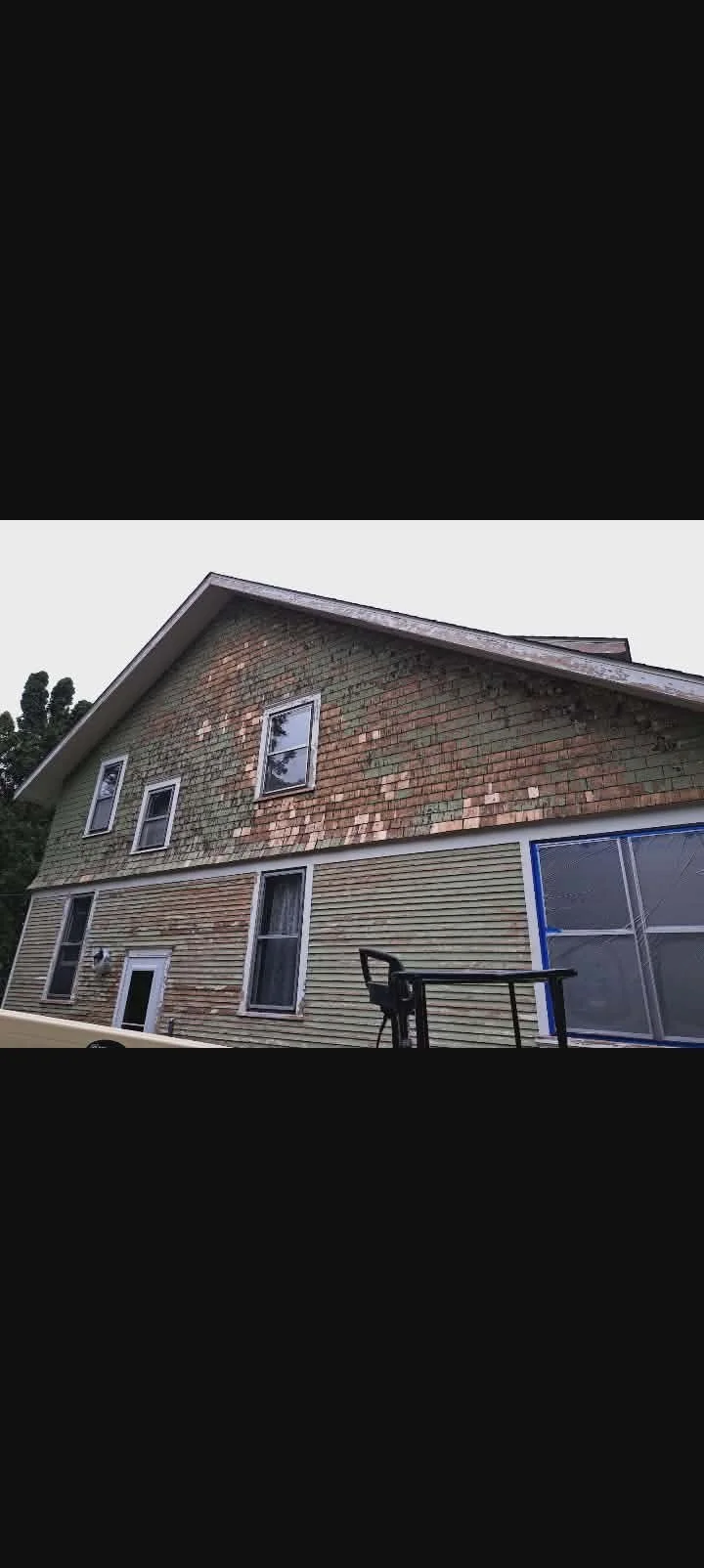 Side view of a two-story house with partially finished exterior, showing a section of old brickwork and new vinyl siding, four windows on the lower level, and three smaller windows on the upper level. There is a backyard with outdoor furniture and tr