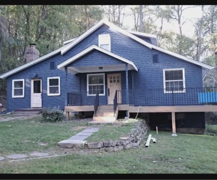 A blue house with a front porch, steps leading up, and multiple windows, surrounded by trees and a grassy yard. After exterior house painting project picture 