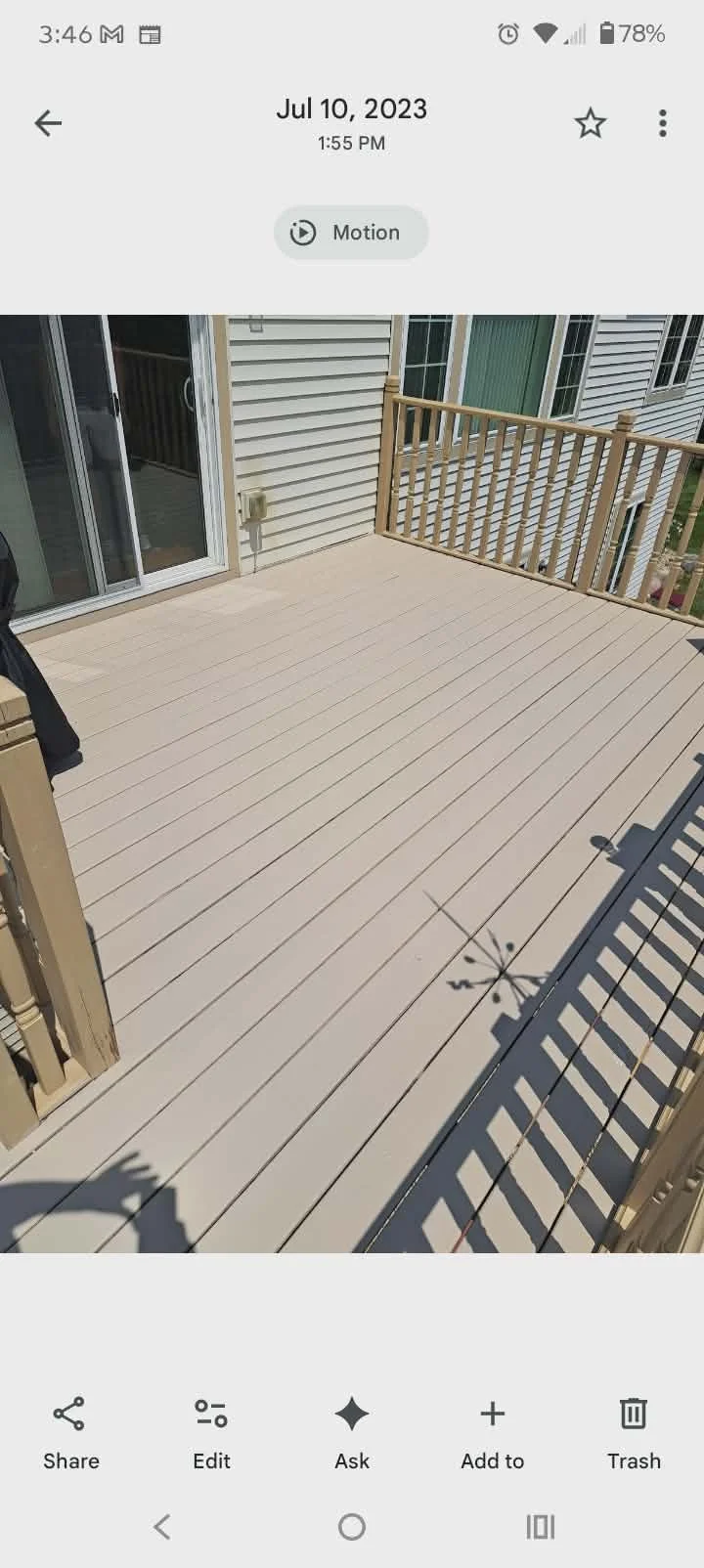 View of a wooden deck outside a house with sliding glass door, beige siding, and a railing, with a shadow of a plant cast on the deck.