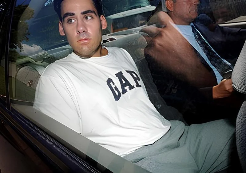 Young man with dark hair and a serious expression sitting in the passenger seat of a car, wearing a white GAP t-shirt, with an older man driving and reading a newspaper.
