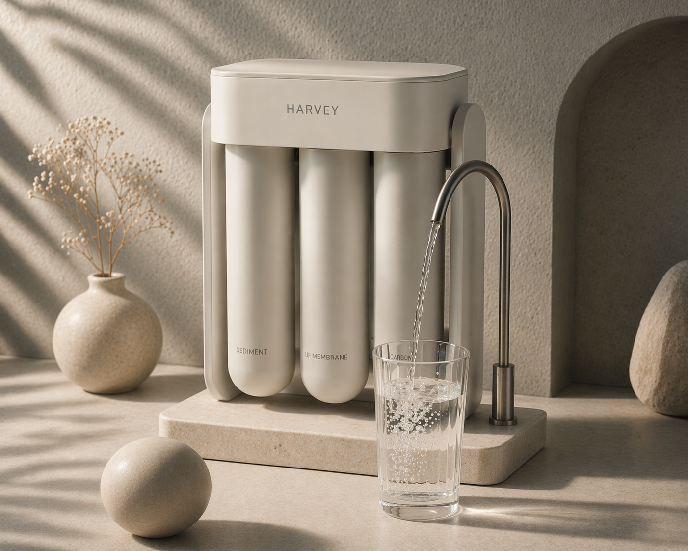 A modern water filtration system with three filter cartridges labeled sediment, UF membrane, and carbon, from the brand Harvey, is shown on a beige countertop with a glass of water being filled from a faucet attached to the system. There is a decorative vase with dried flowers and a round stone beside it in a sunlit room.