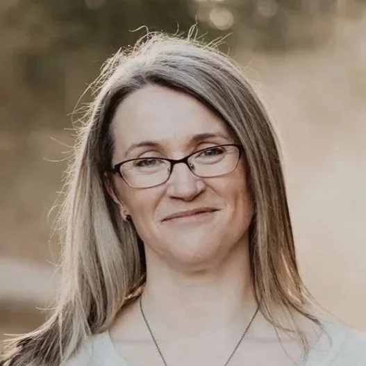 A woman with glasses and long hair smiling outdoors in natural light.