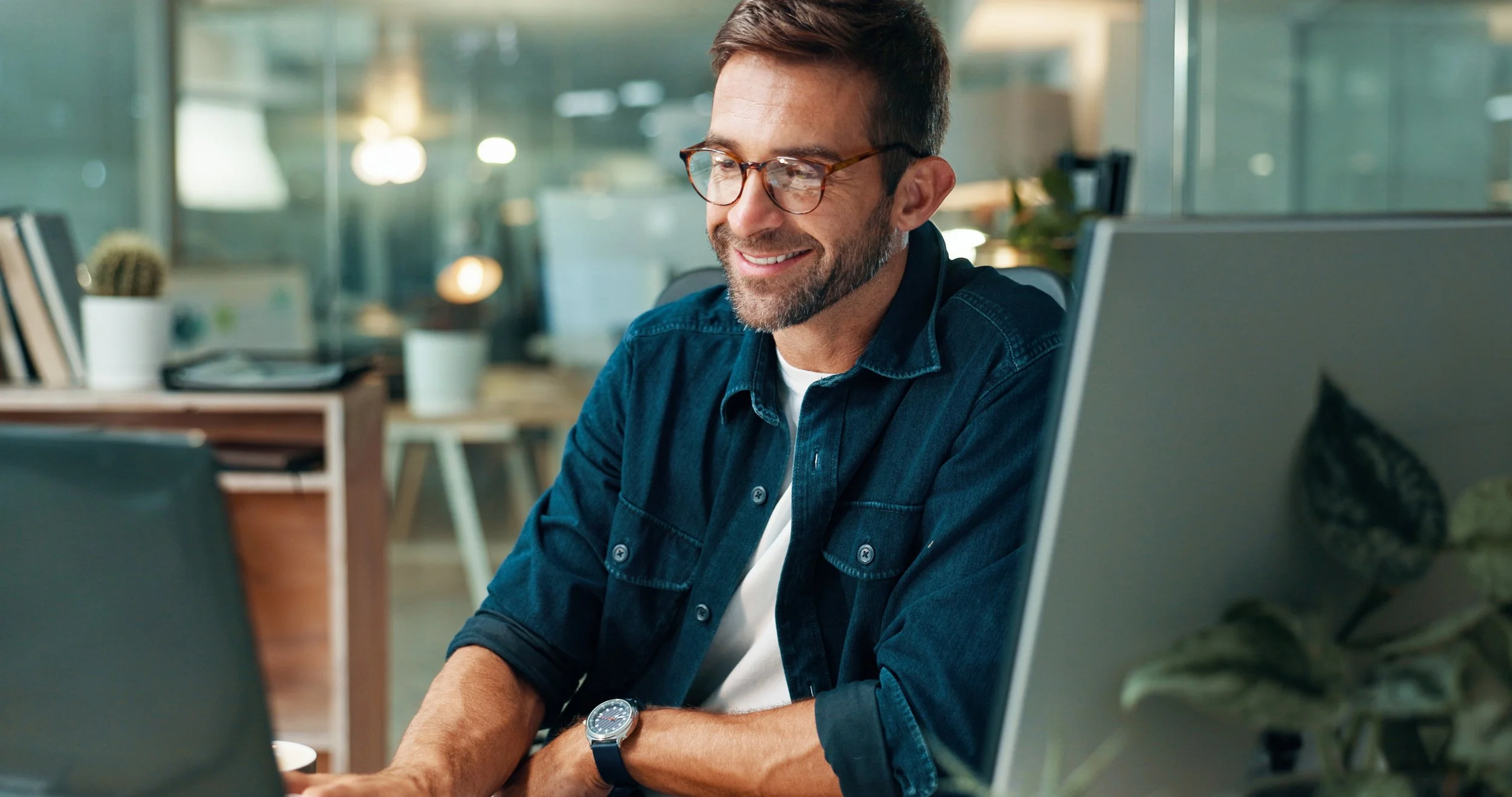 A smiling man with glasses and a beard, wearing a denim shirt, working on a laptop in a modern home office.