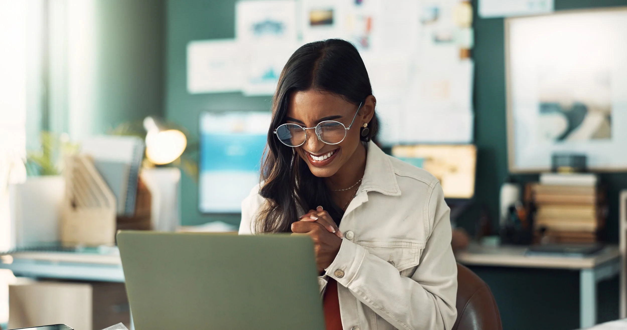A woman with long dark hair and pale brown skin, wearing glasses, earrings and a white jacket. She is looking at a laptop and smiling happily.
