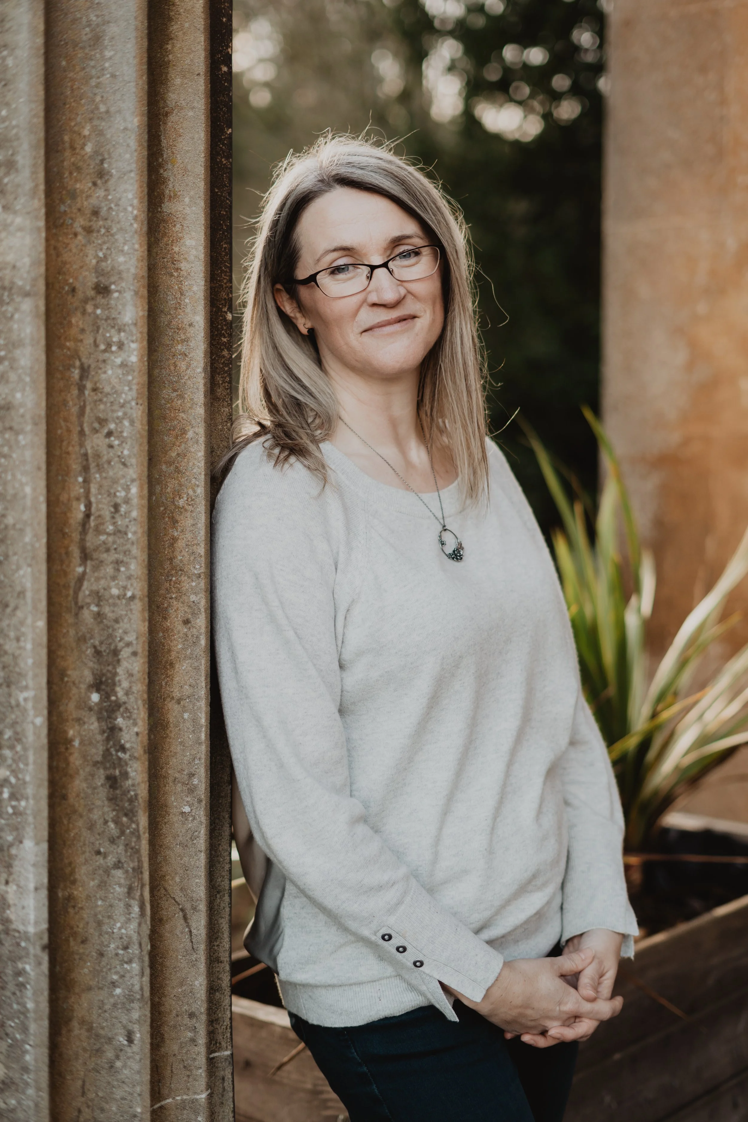A smiling woman with glasses and shoulder-length hair leaning against an outdoor wall, with greenery in the background, wearing a light grey sweater and dark jeans.