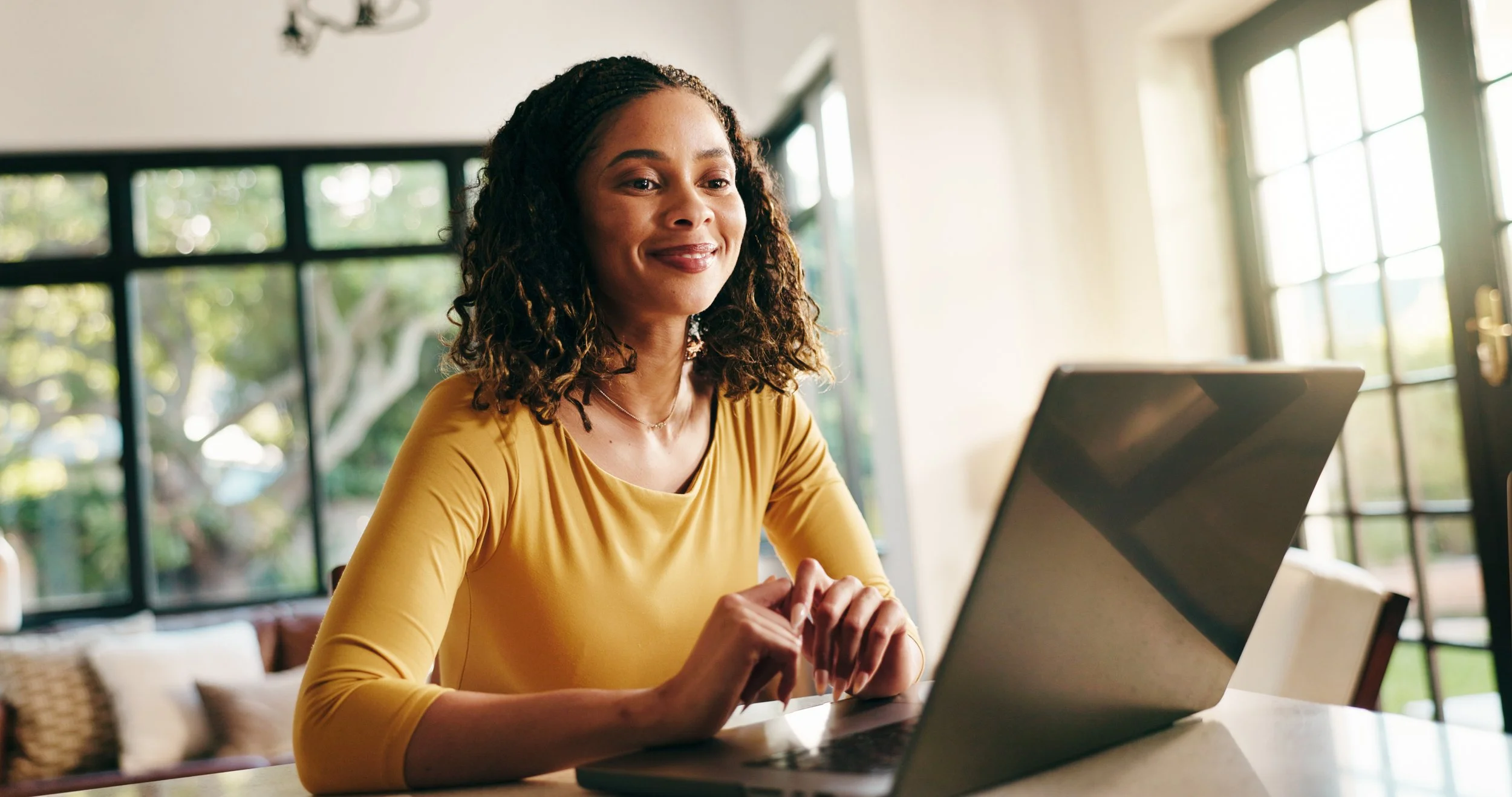 A woman with dark curly hair and a yellow shirt smiling with satisfaction while working on a laptop in a bright room with large windows.