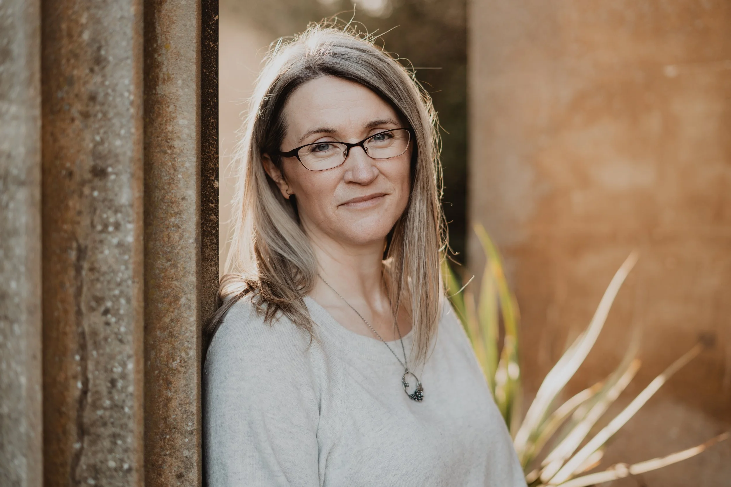 A smiling woman with glasses and long hair standing outdoors near a sandstone wall, with plants and sunlight in the background.