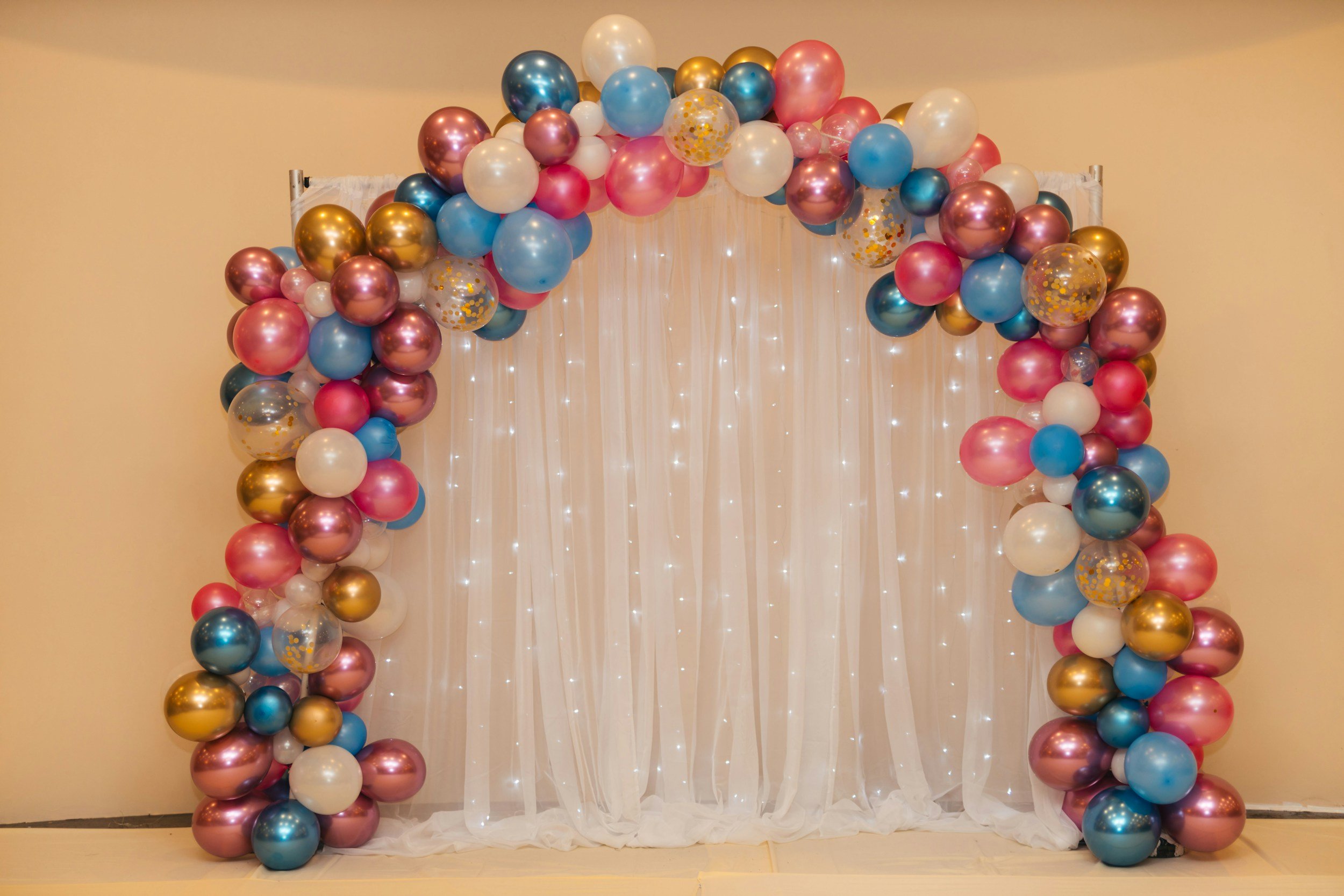 A balloon arch with various colors including pink, blue, white, gold, and confetti-filled balloons, set against a cream-colored backdrop with hanging string lights.