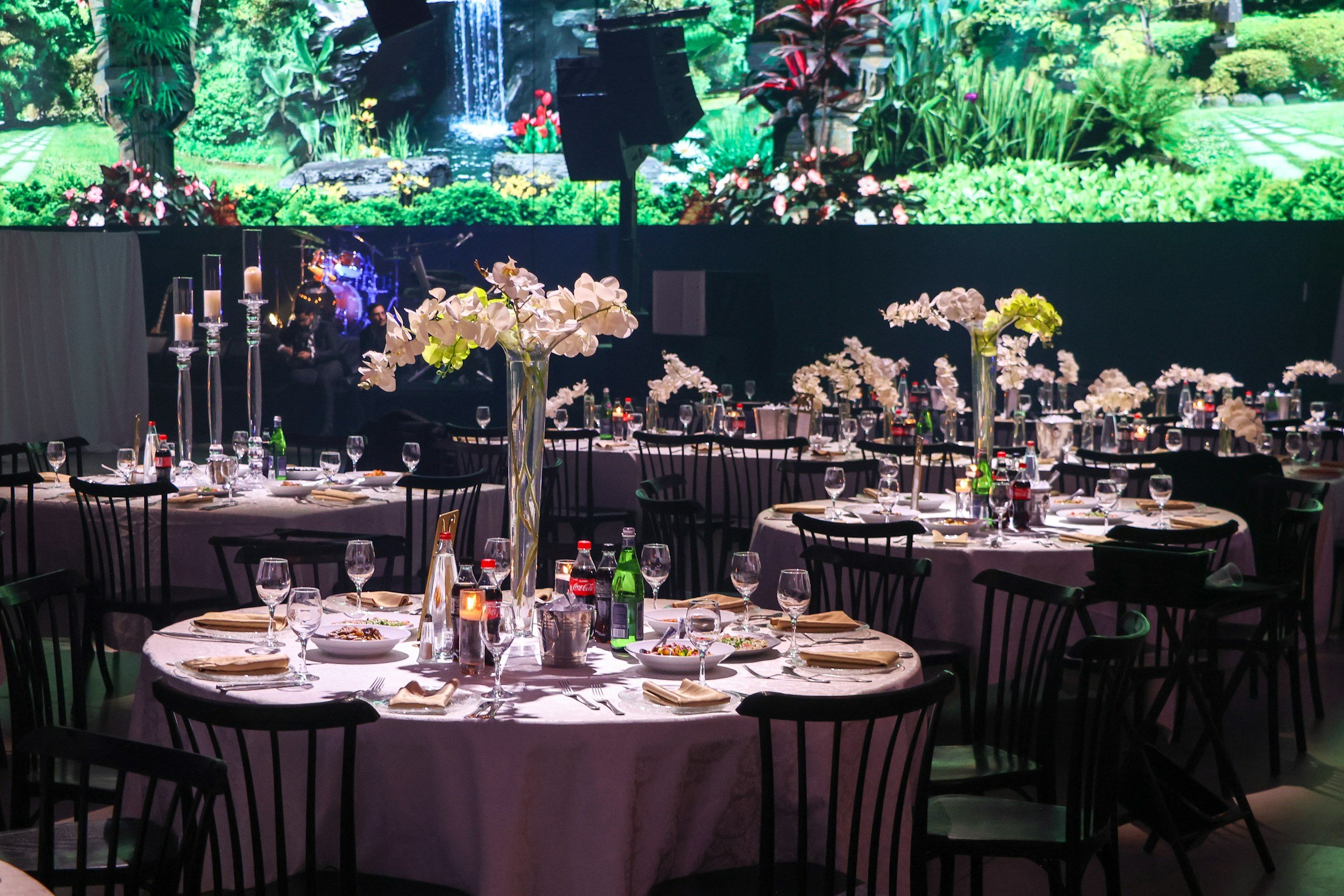 Round banquet tables with place settings, floral centerpieces, and drinks in a banquet hall. A large digital screen behind displays a lush garden scene with a waterfall and greenery.