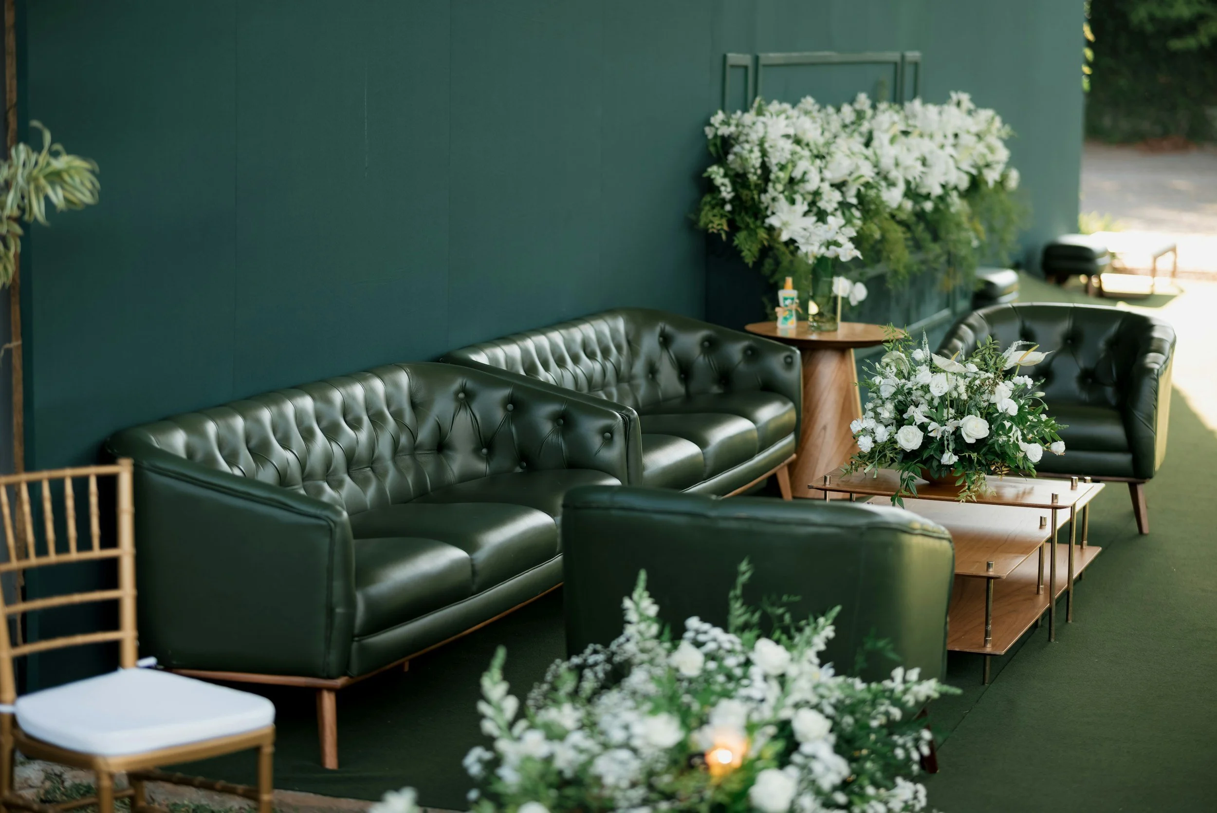 An outdoor seating area with green leather tufted couches, a wooden table decorated with a large floral arrangement, and a dark green wall background.