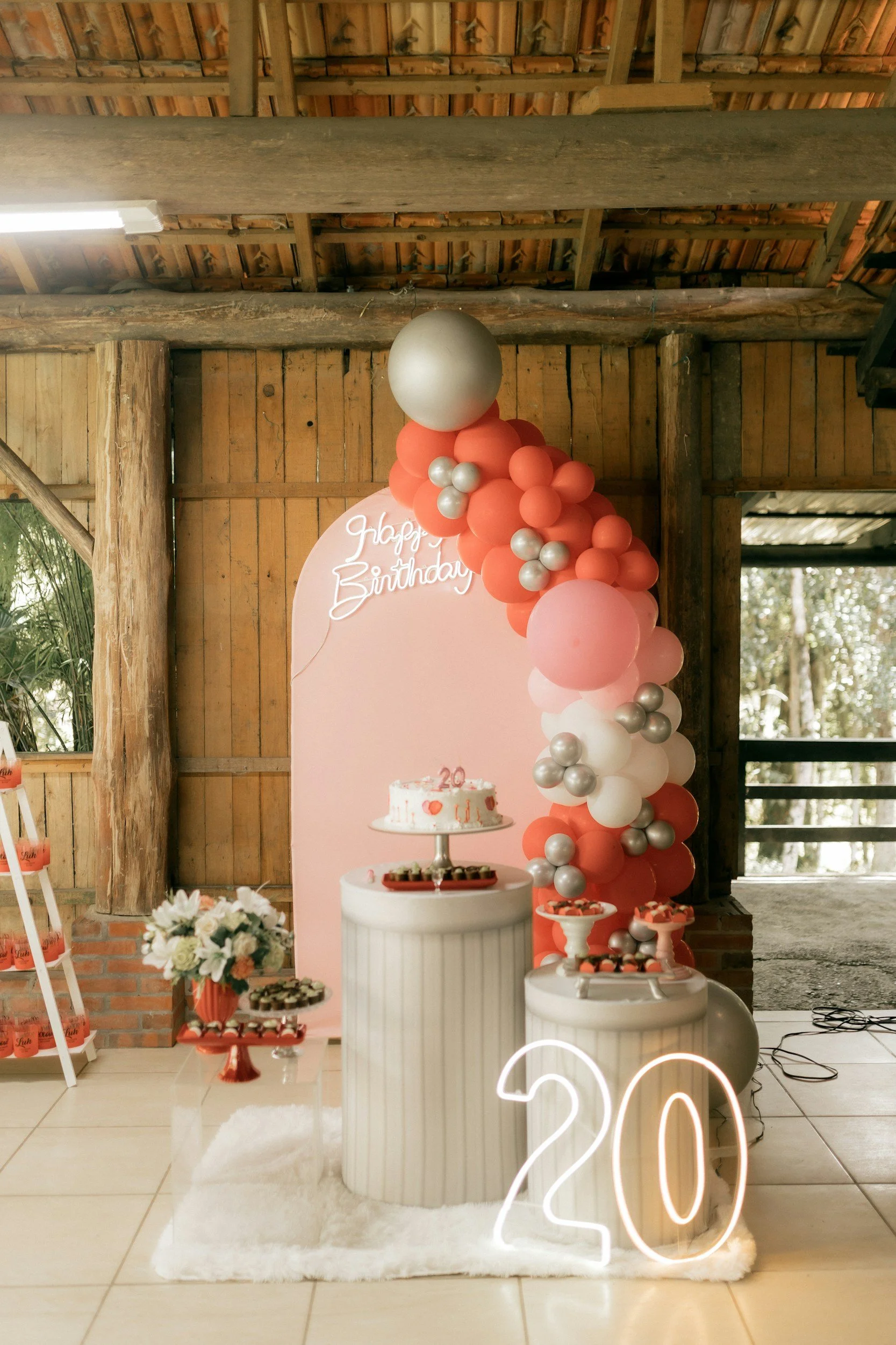 Decorative birthday celebration with pink, peach, white, and silver balloons arranged in an arch, a pink backdrop with a neon sign that says 'Happy Birthday,' a table with a cake and cupcakes, floral arrangements, and a large illuminated '20' sign on the floor. The setting is in a rustic wooden venue with natural wood beams and walls.