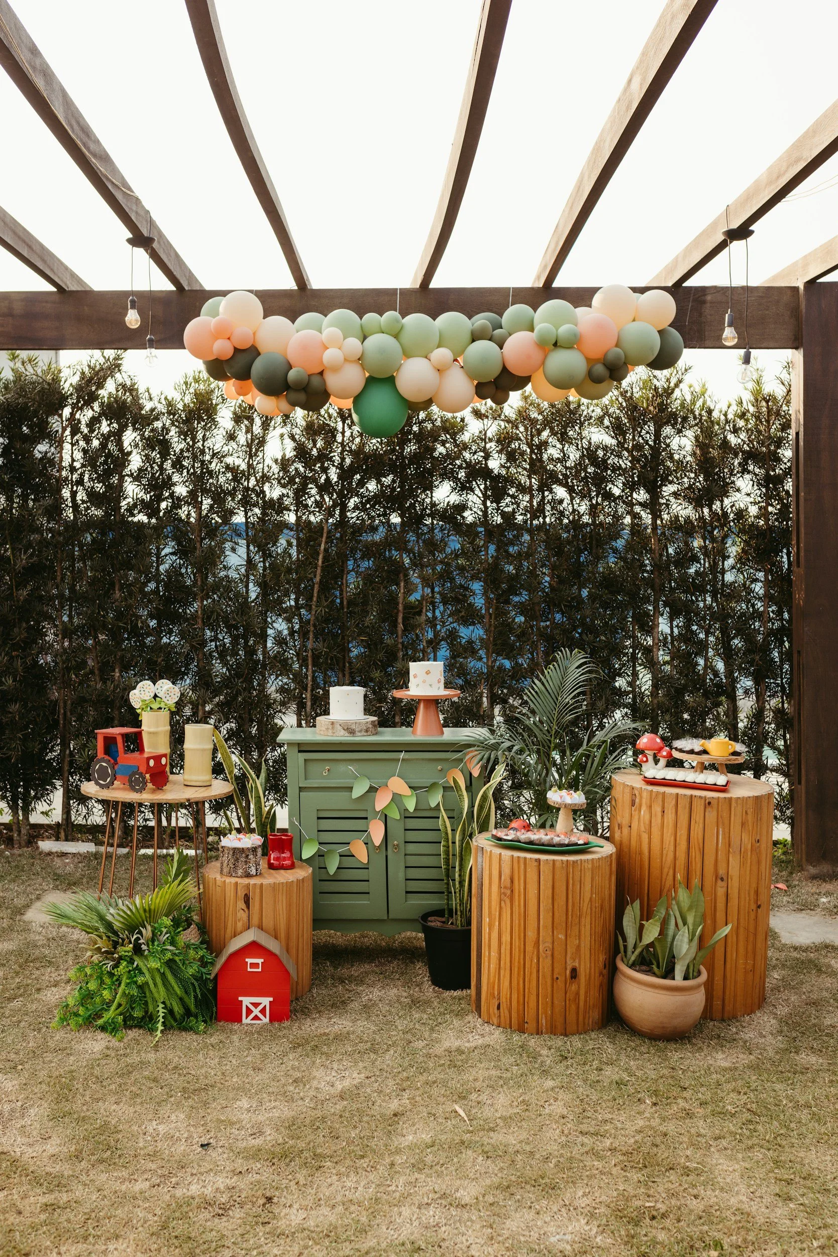 Party setup with a balloon garland, decorated tables with cakes, plants, and wooden accents, outdoors with a wooden pergola and string lights.