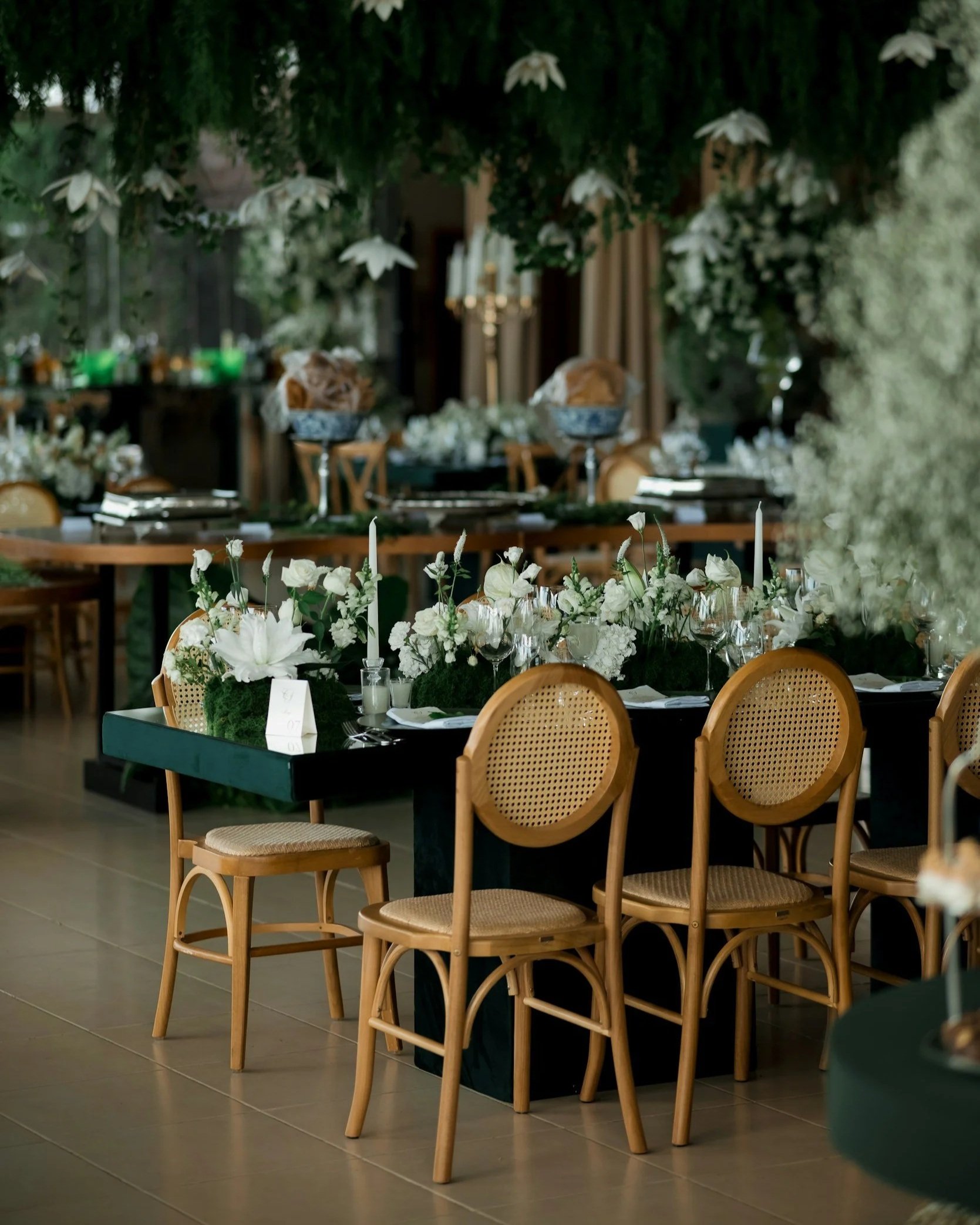 Indoor wedding reception setup with round tables decorated with white floral centerpieces, surrounded by wooden chairs with cane backs, and draped greenery and flowers hanging from the ceiling.