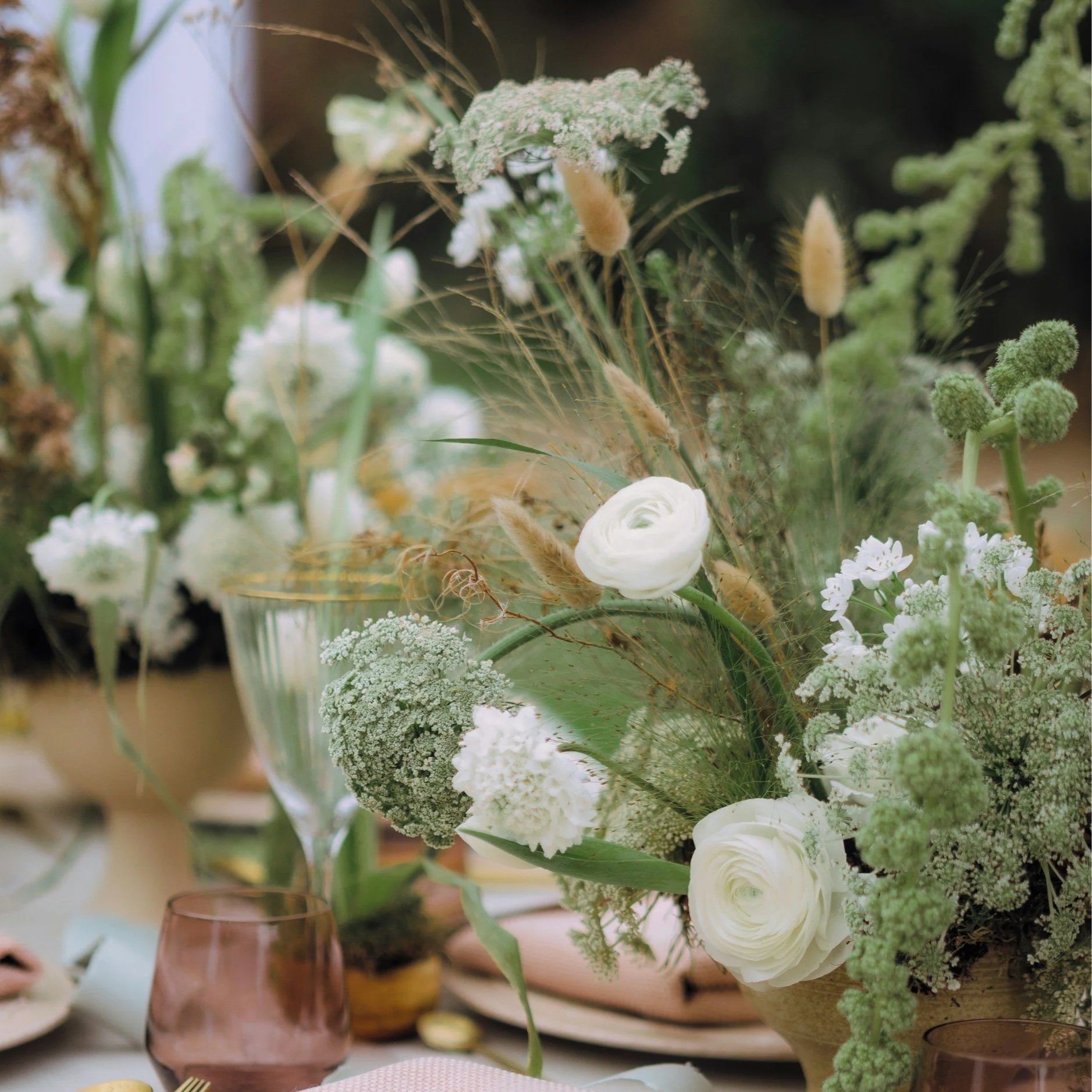 A floral arrangement with white and green flowers in a brown vase on a table, with other vases and flowers in the background.
