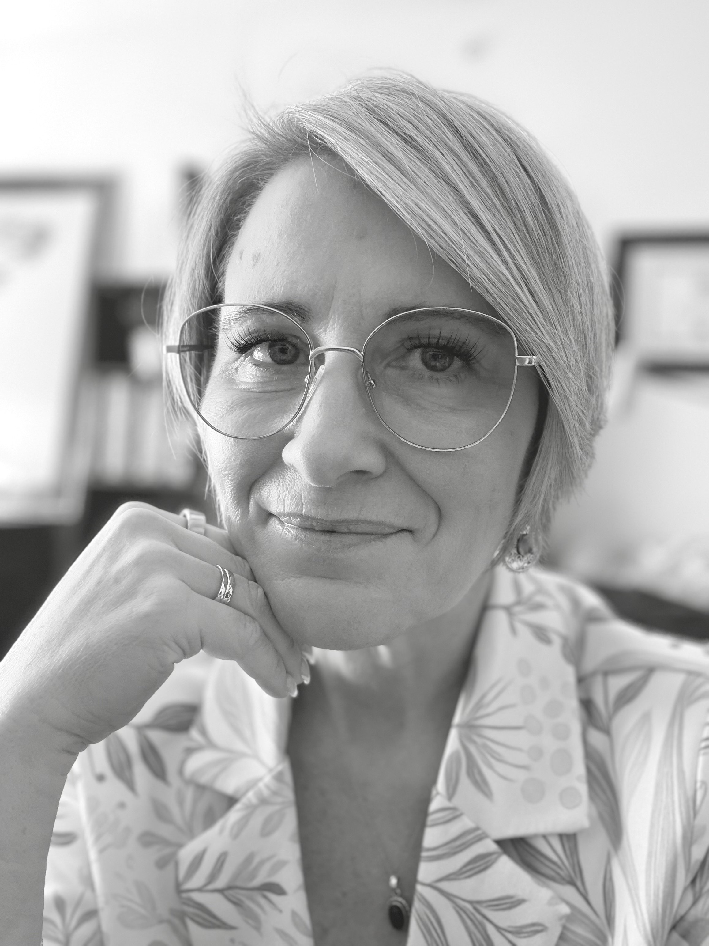A woman with short, light-colored hair wearing glasses and a patterned blouse, smiling gently and resting her chin on her hand.