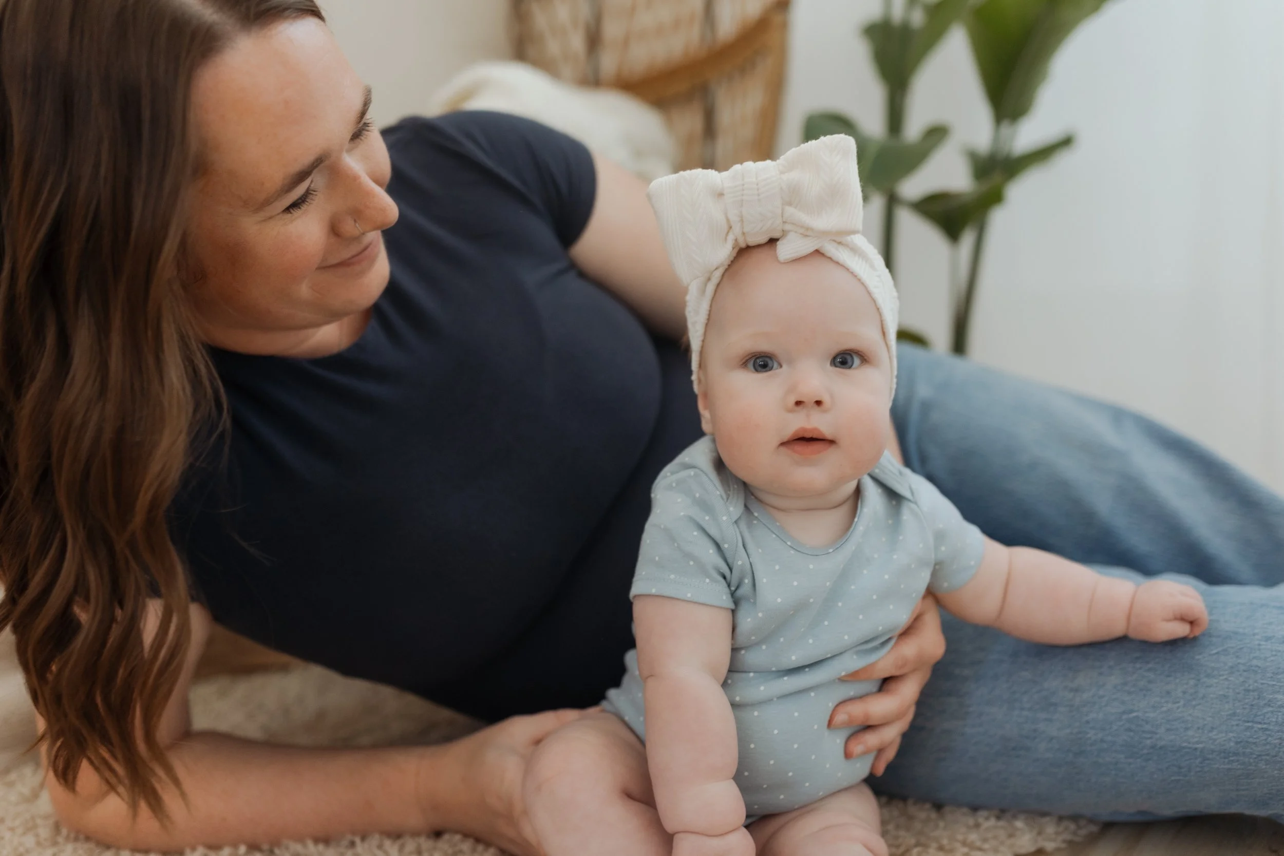 A woman with long red hair and a nose piercing is smiling and playing with a baby. The baby, sitting on the woman's lap, has blue eyes and is wearing a gray shirt with white polka dots and a large cream-colored bow headband. The background includes a wicker chair, some greenery, and a white wall.