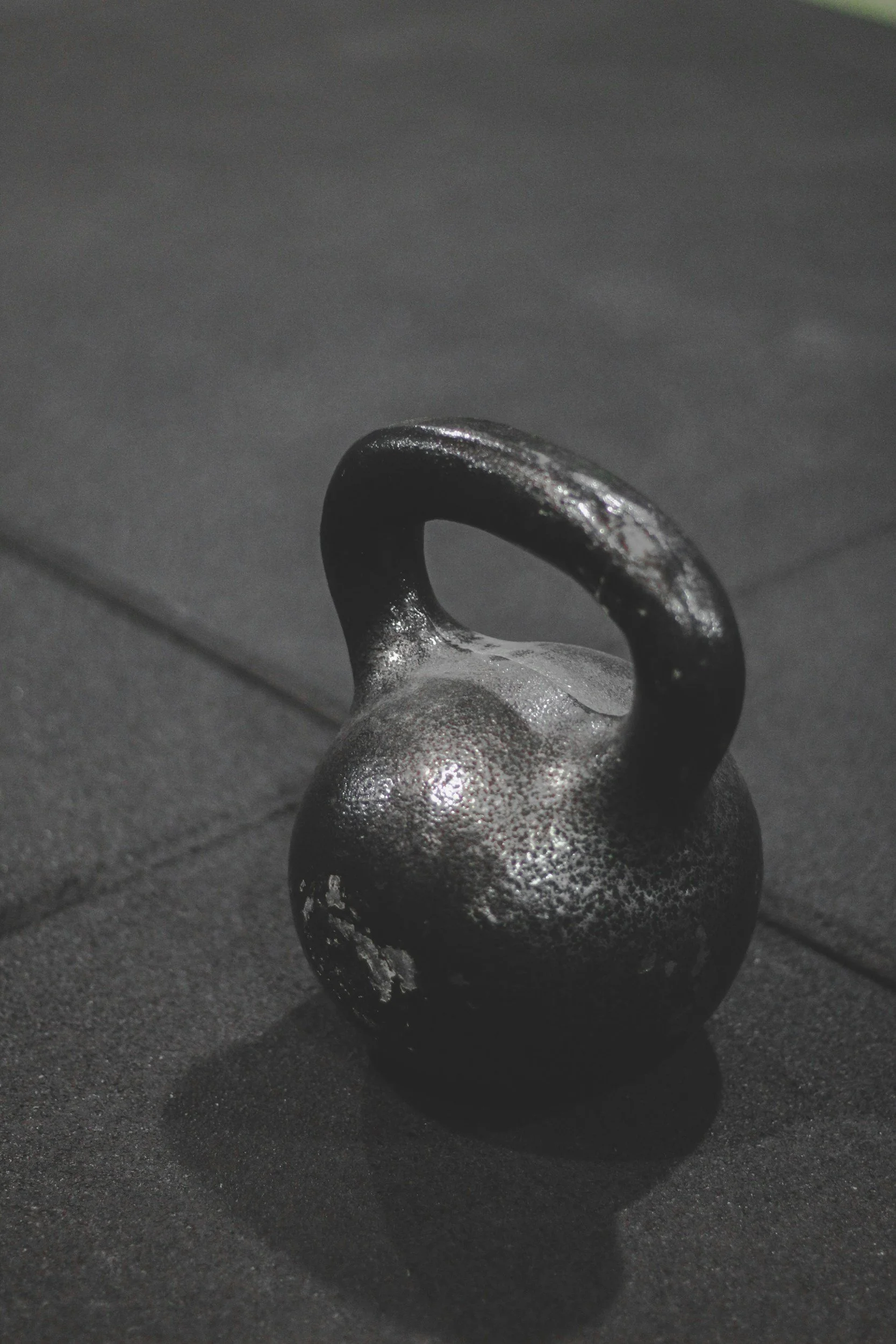 A close-up photo of a metal kettlebell on a dark surface, with its handle prominently visible and casting a shadow.