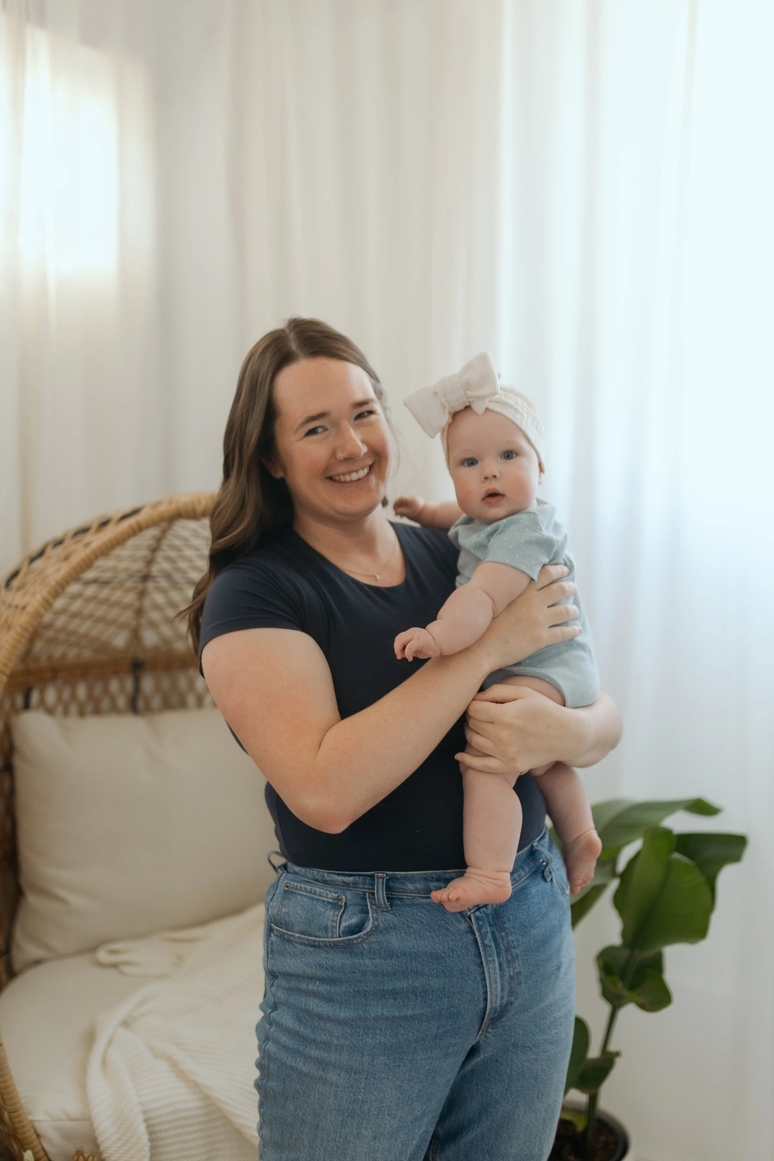 A woman holding a baby girl indoors near a white curtain and green plant wall.