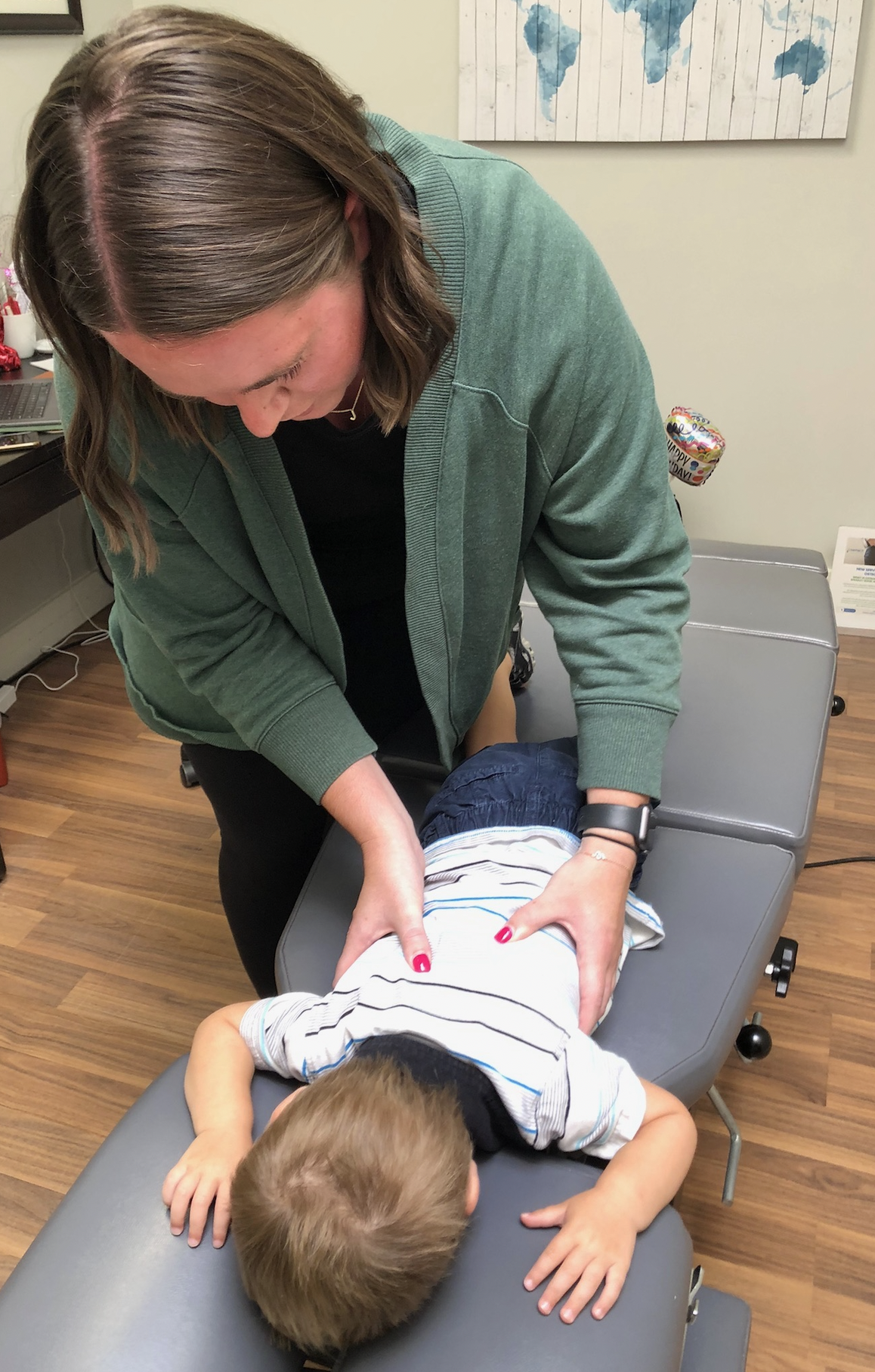 A woman providing chiropractic care or spinal adjustments to a young boy lying face down on a treatment table in a medical office. The woman is leaning over and applying pressure with her hands on the boy's back. There is a world map poster on the wall behind them, and a small balloon with