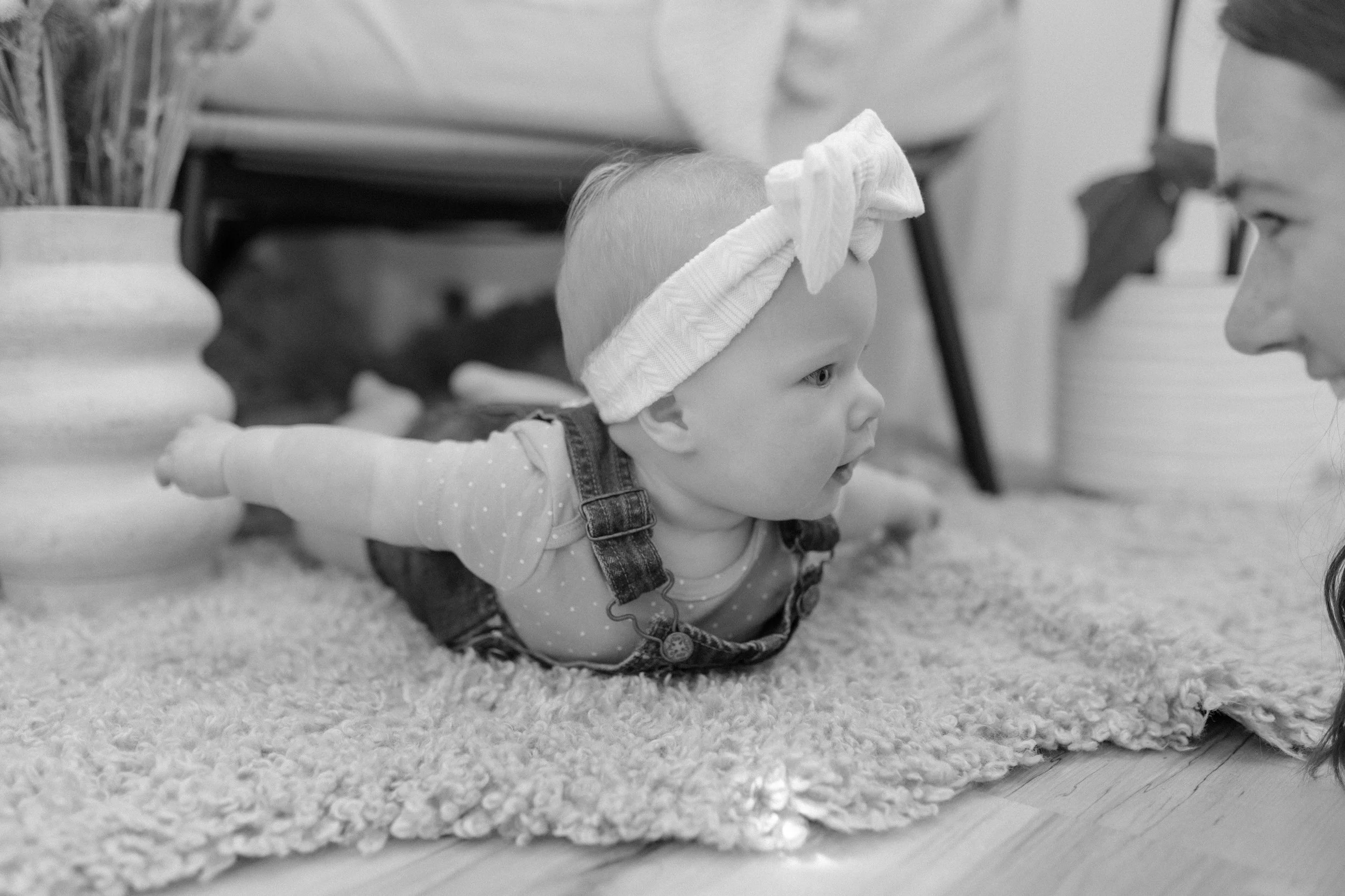 A young child with light hair and wearing overalls and a headband is crawling on a carpeted floor, reaching out with one arm and looking towards a person whose face is partially visible. The scene is in black and white.
