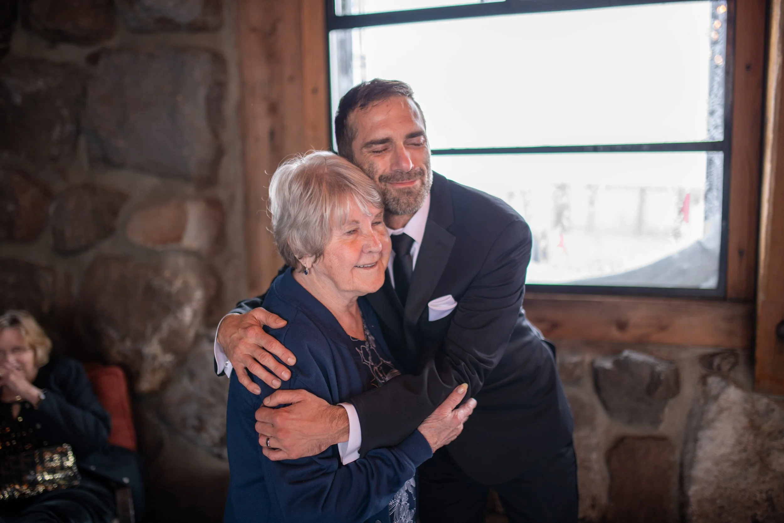 A man and an older woman hugging each other, smiling, in a cozy room with a stone wall and large window in the background