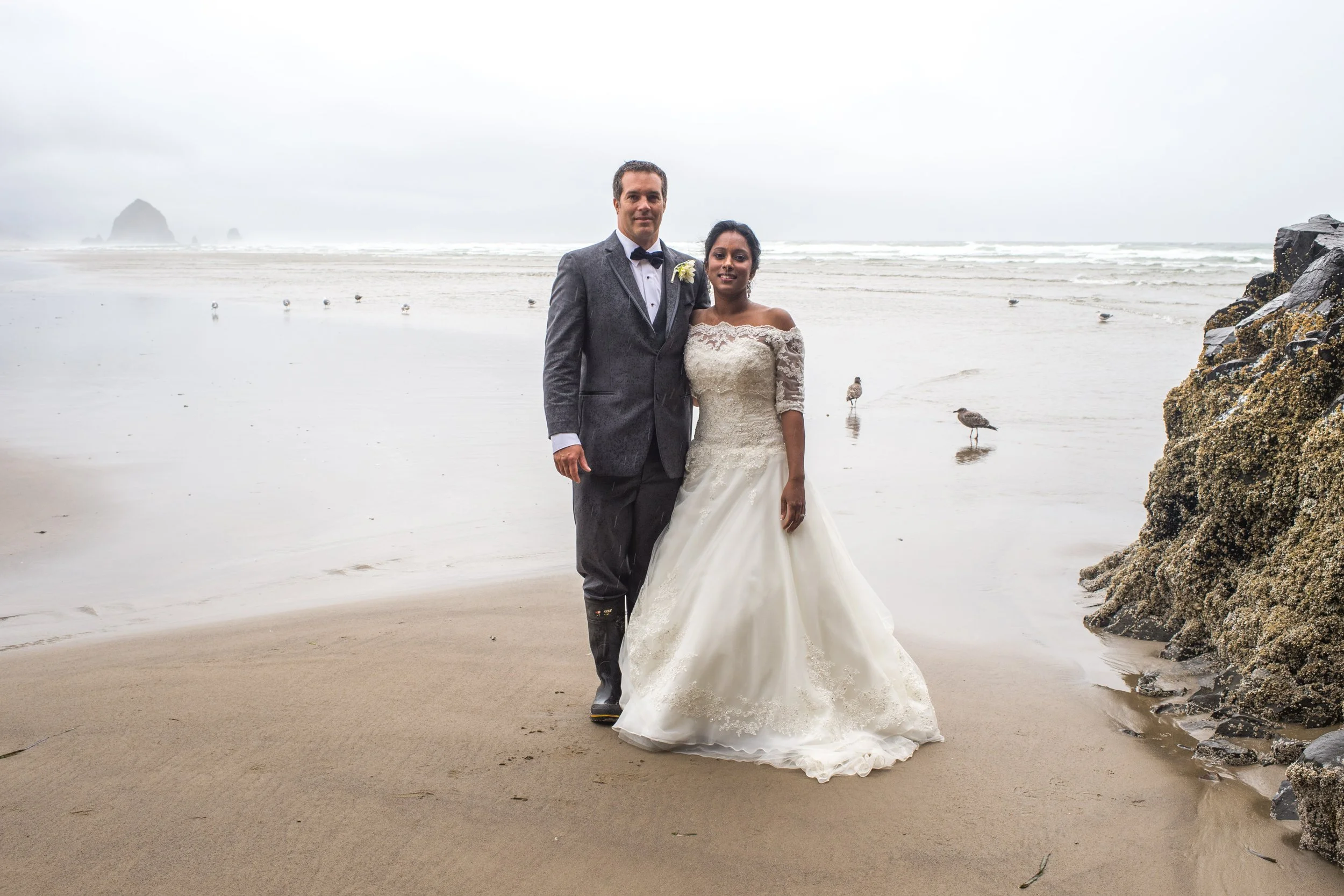A newlywed couple standing on the beach, with the groom in a gray suit and bow tie, and the bride in a white lace wedding dress, near rocks and seagulls, with an ocean and rocky island in the background under overcast sky.