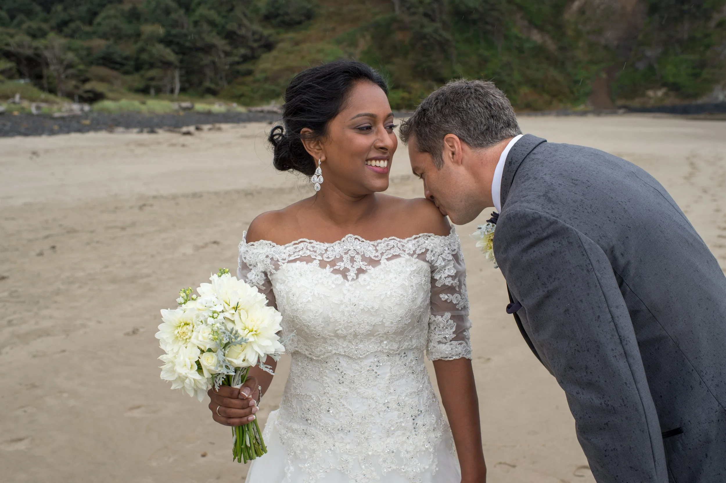 A bride in a lace wedding gown holding a bouquet of white flowers, smiling as a groom kisses her on the forehead on a beach with green hills in the background.