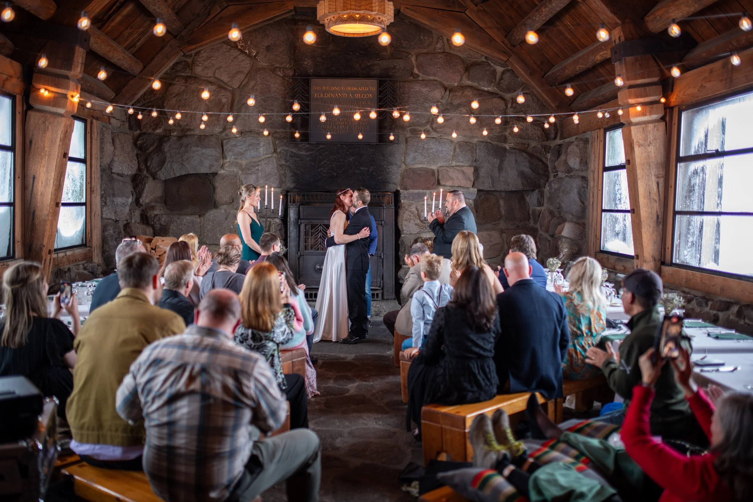 A wedding ceremony inside a rustic stone and wood venue with string lights hanging from the ceiling. The bride and groom are kissing in front of an audience seated at tables, with a priest or officiant on the side. The room has large windows and wood