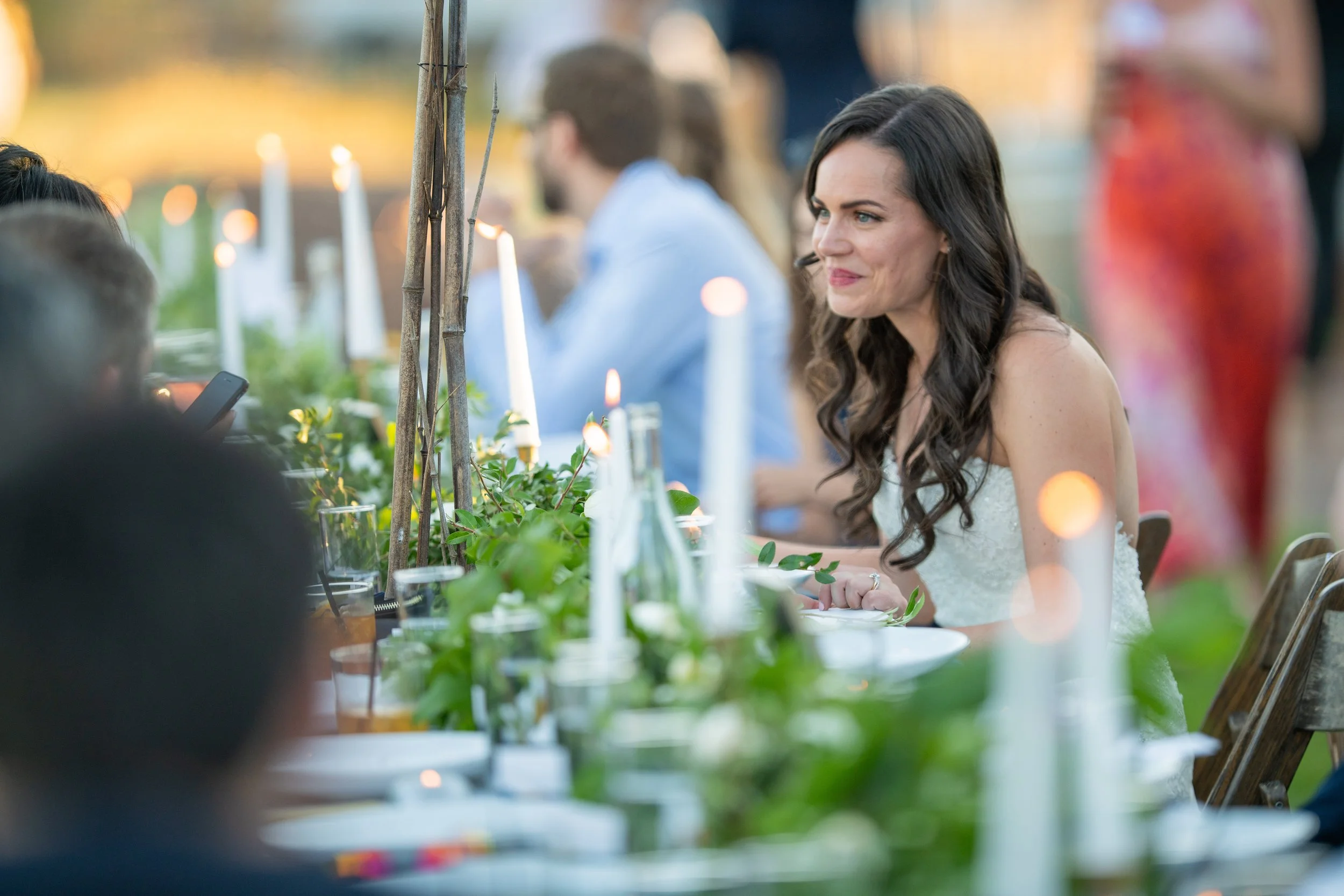 A woman in a wedding dress sitting at a decorated outdoor reception table with candles and greenery, smiling and looking to her right.