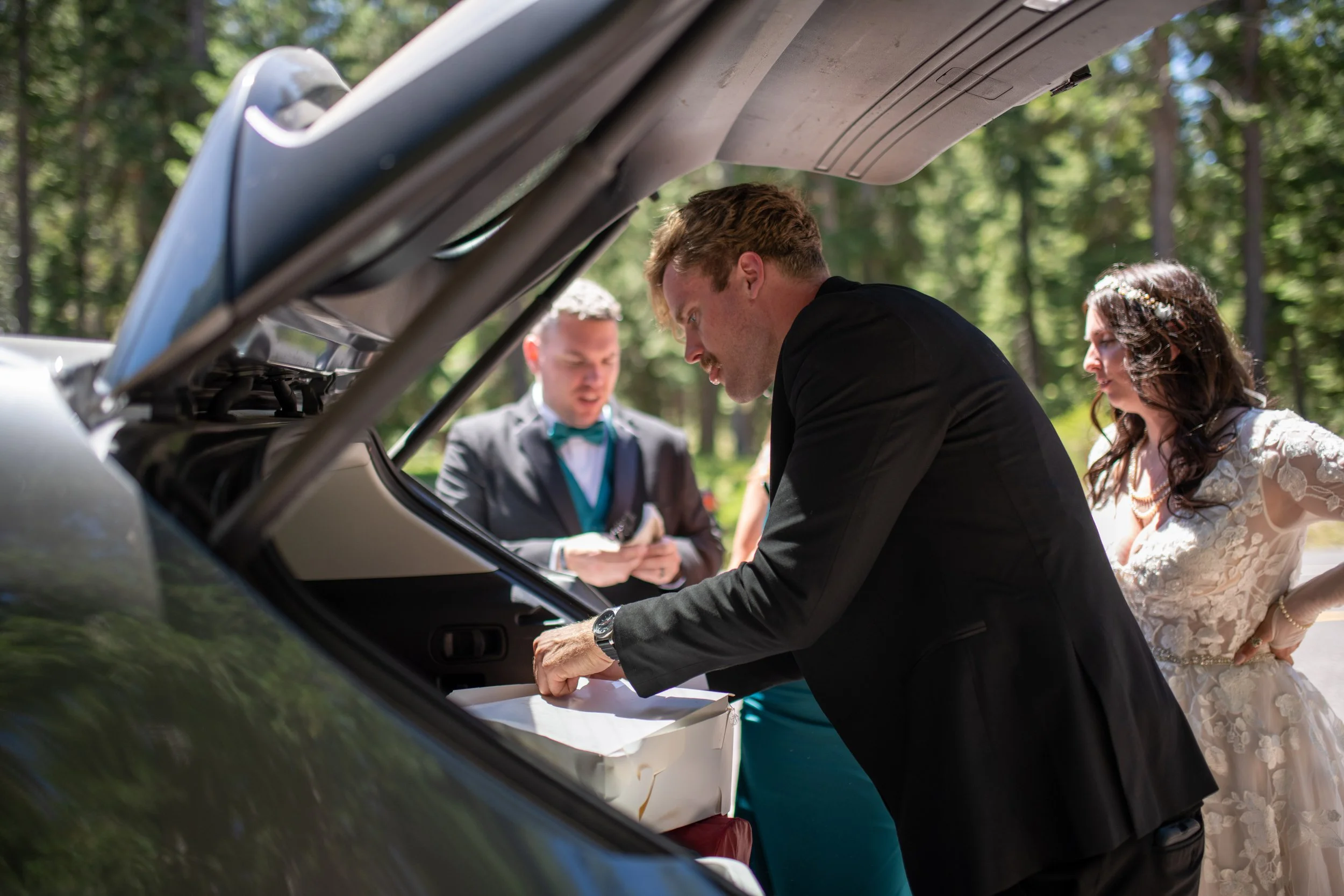 A wedding ceremony with a bride and groom signing paperwork in a car trunk, with a man in a gray suit and tie and a woman in a white lace dress standing outside in a wooded area.