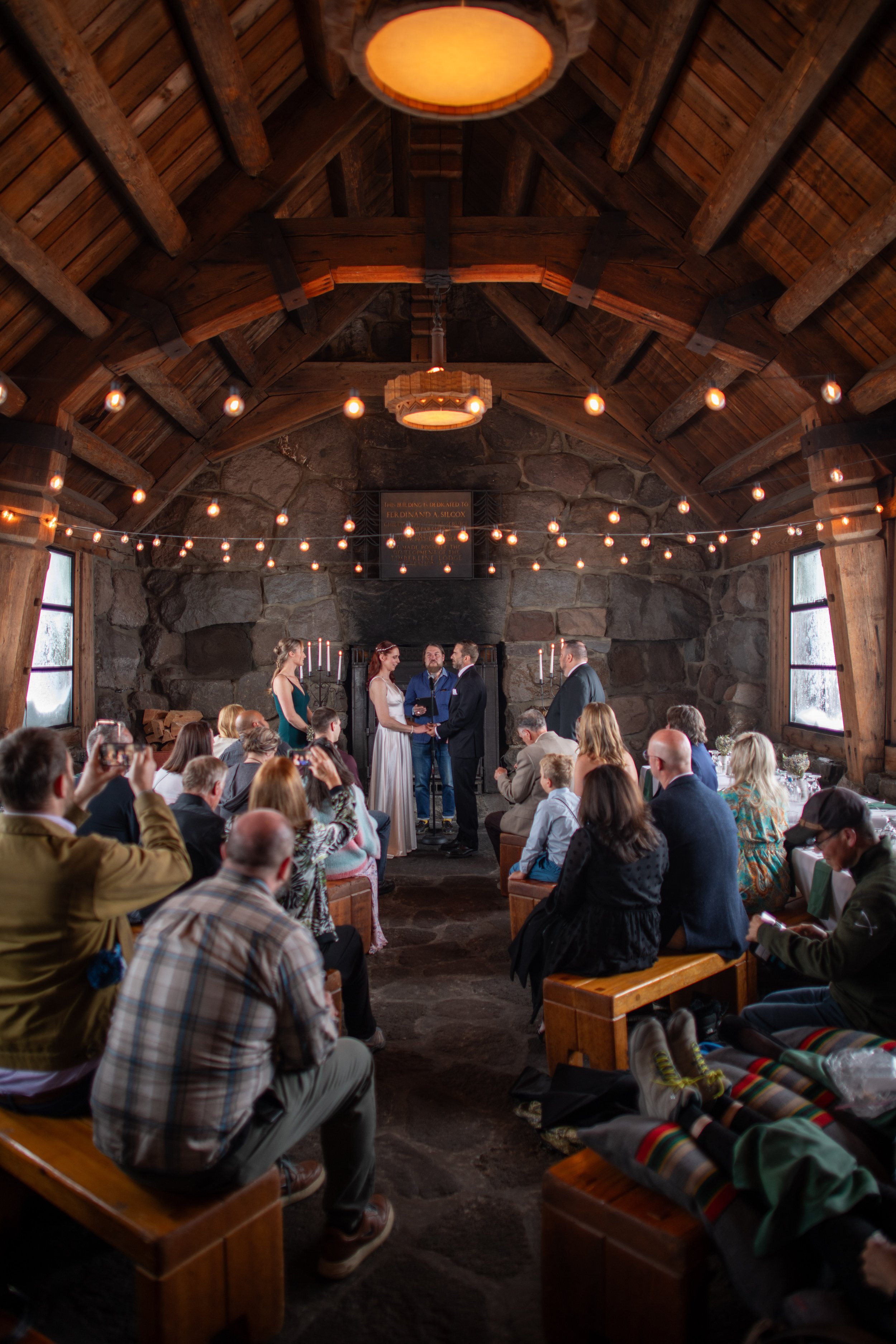 A wedding ceremony inside a rustic wooden chapel with string lights and a stone wall backdrop. The bride and groom are facing each other with an officiant, while guests are seated watching.